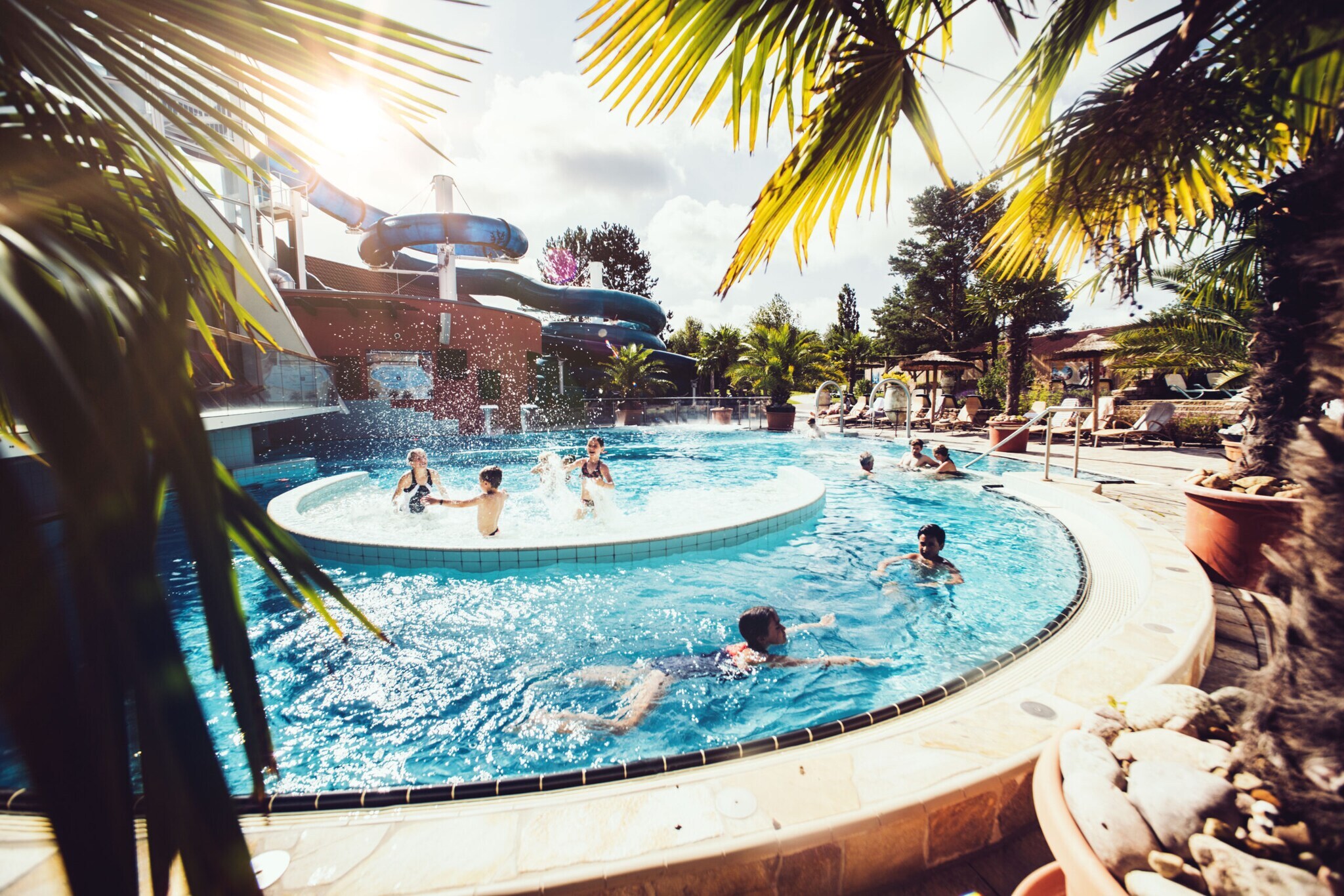 Badelandschaft mit Kindern im Außenpool vor einem Komplex mit Baderutsche, im Vordergrund Palmen