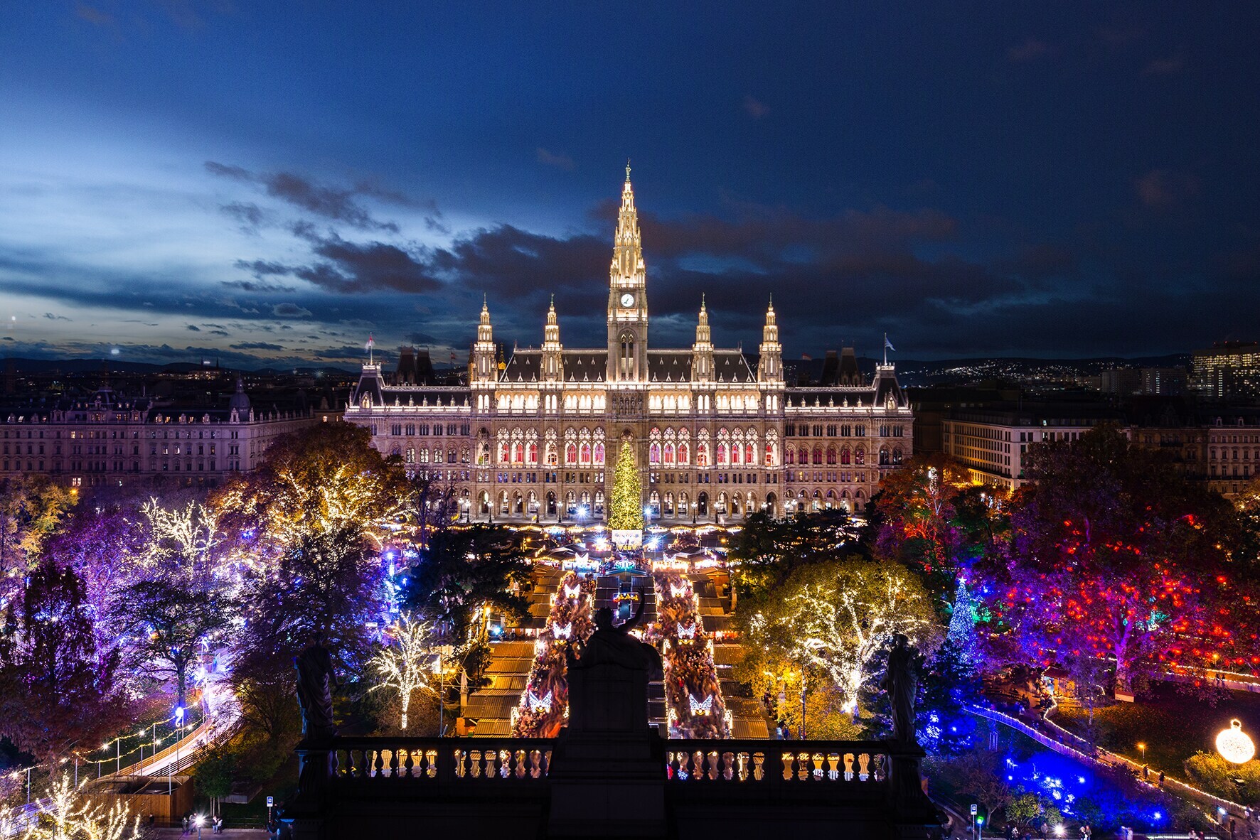 Panoramablick auf das hell erleuchtete Wiener Rathaus und den Rathausvorplatz mit bunt beleuchteten Bäumen bei Nacht Panoramablick auf das hell erleuchtete Wiener Rathaus und den Rathausvorplatz mit bunt beleuchteten Bäumen bei Nacht