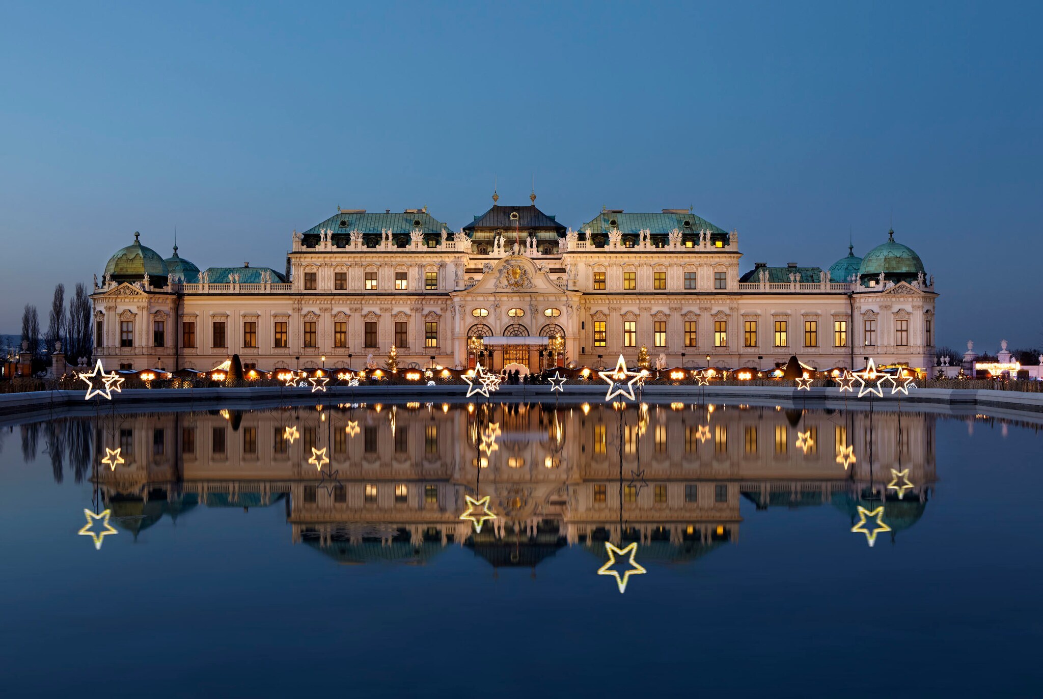 Das erleuchtete Wiener Barockschloss Belvedere mit Weihnachtsmarkt spiegelt sich in der Wasseroberfläche in der Abenddämmerung