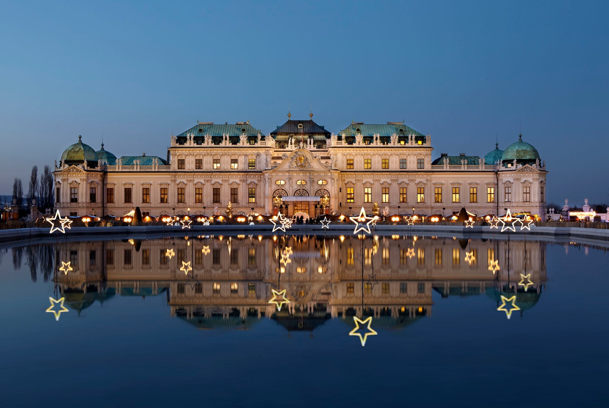 Das erleuchtete Wiener Barockschloss Belvedere mit Weihnachtsmarkt spiegelt sich in der Wasseroberfläche in der Abenddämmerung Das erleuchtete Wiener Barockschloss Belvedere mit Weihnachtsmarkt spiegelt sich in der Wasseroberfläche in der Abenddämmerung