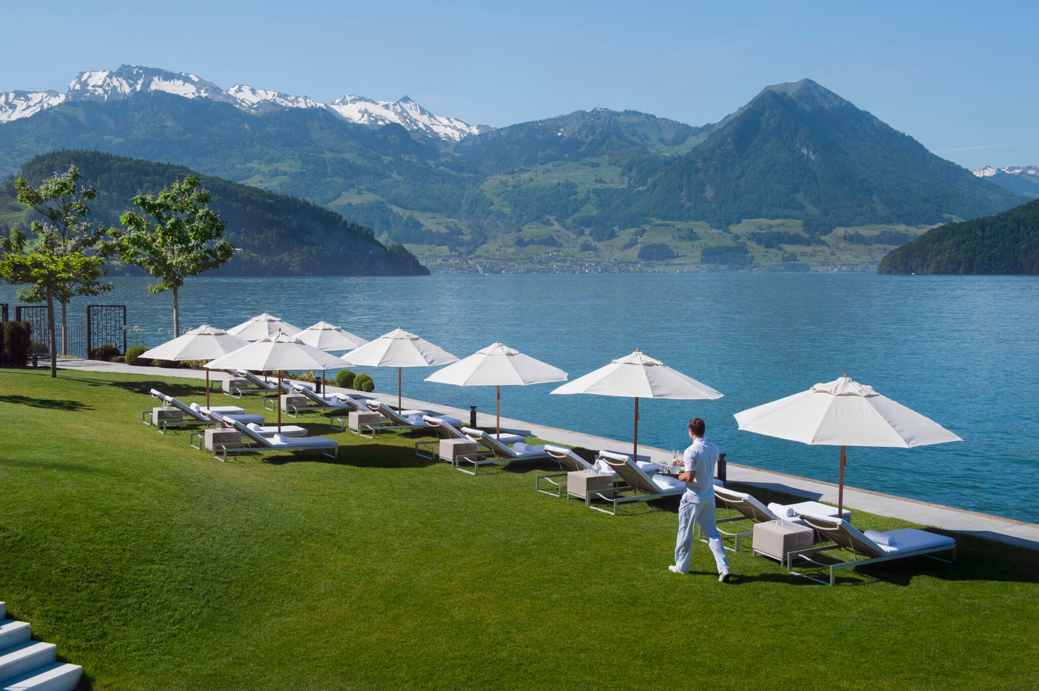 Badeliegen mit Sonnenschirmen auf einer Wiese am Seeufer, im Hintergrund Bergpanorama