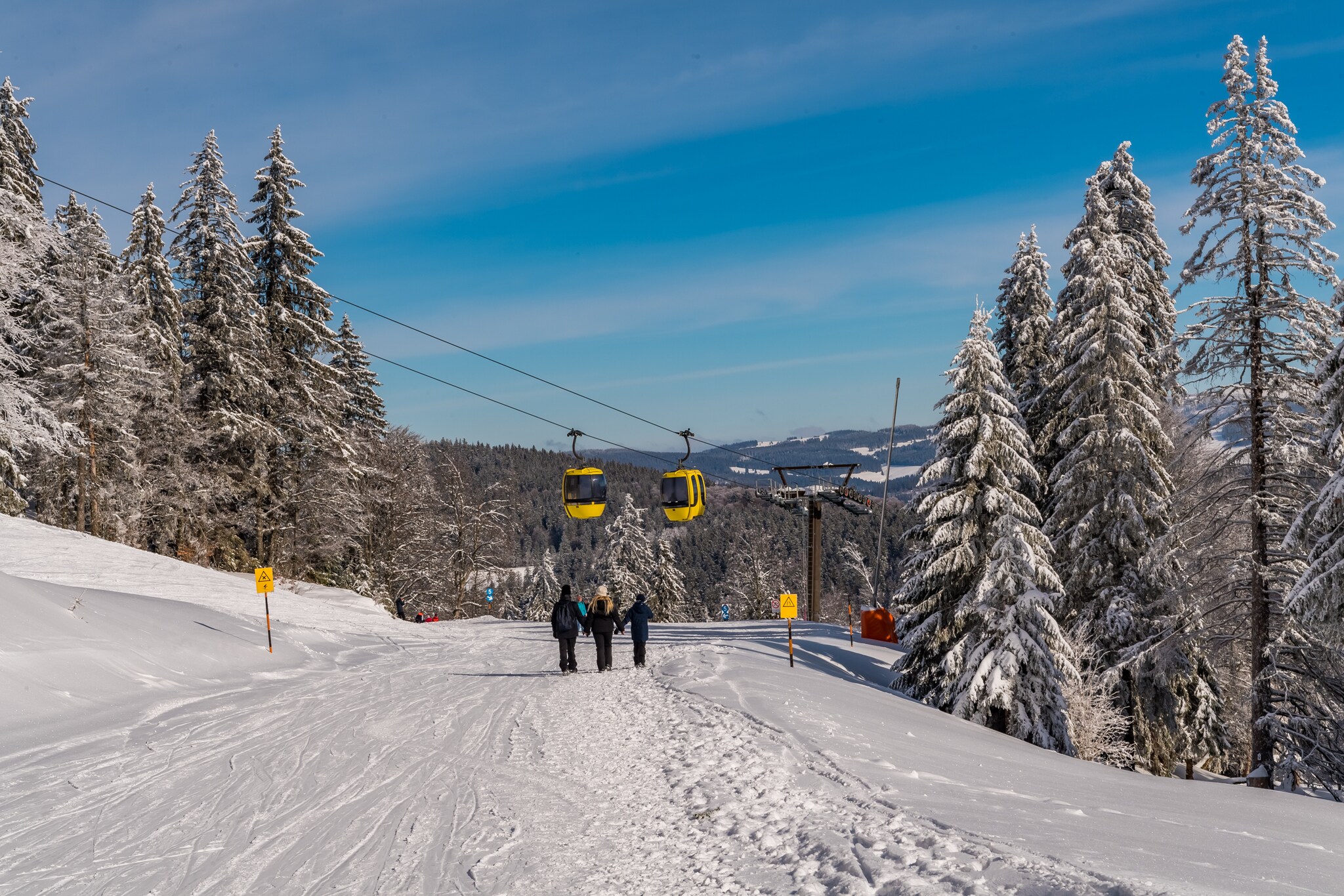 Drei Personen gehen eine Skipiste hinab, über ihnen fahren gelbe Gondeln hinweg