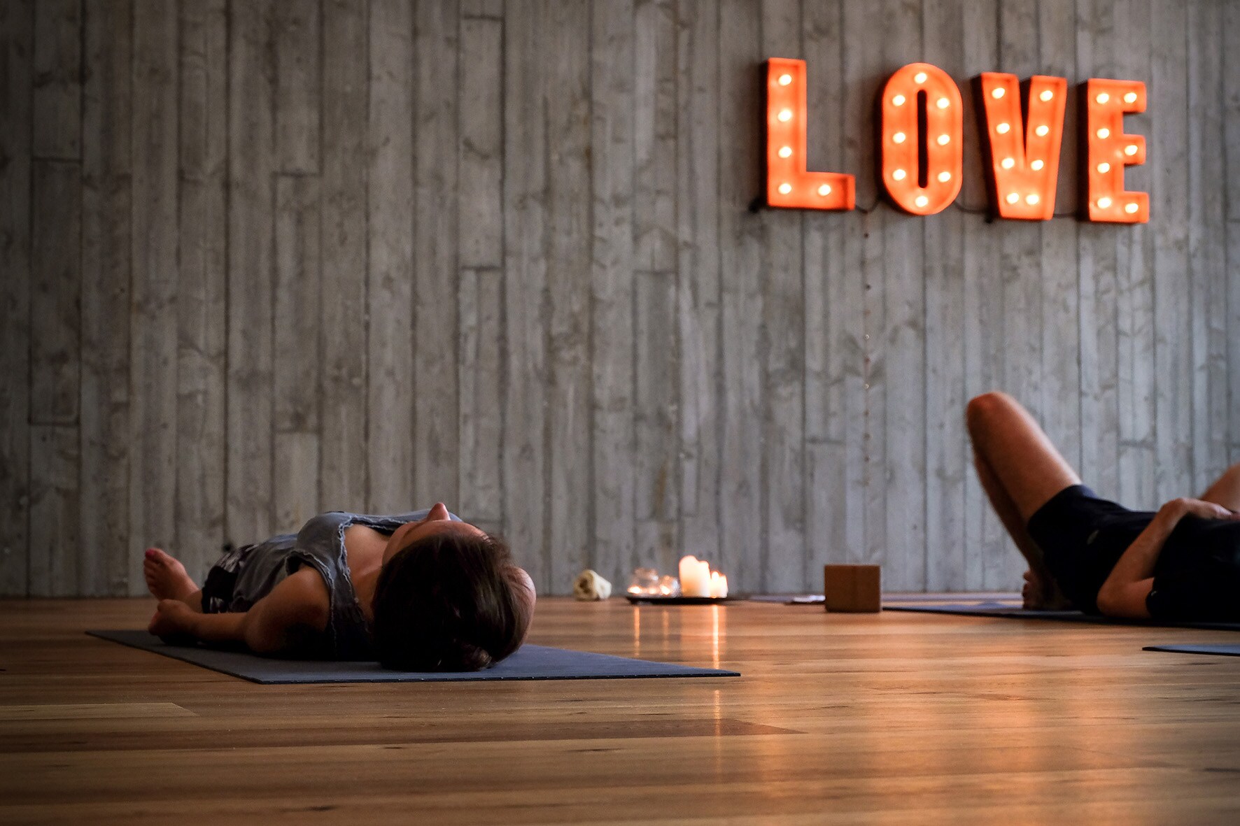Zwei Personen liegen in einem gemütlichen Yoga-Raum auf ihren Matten, auf dem Boden stehen Kerzen und an der Wand leuchten die Lettern LOVE Zwei Personen liegen in einem gemütlichen Yoga-Raum auf ihren Matten, auf dem Boden stehen Kerzen und an der Wand leuchten die Lettern LOVE
