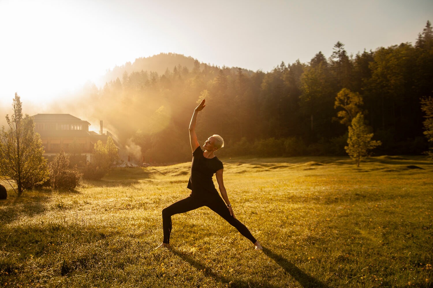 Eine Frau macht Yoga bei Sonnenaufgang und noch leichtem Nebel in einem grünen Garten