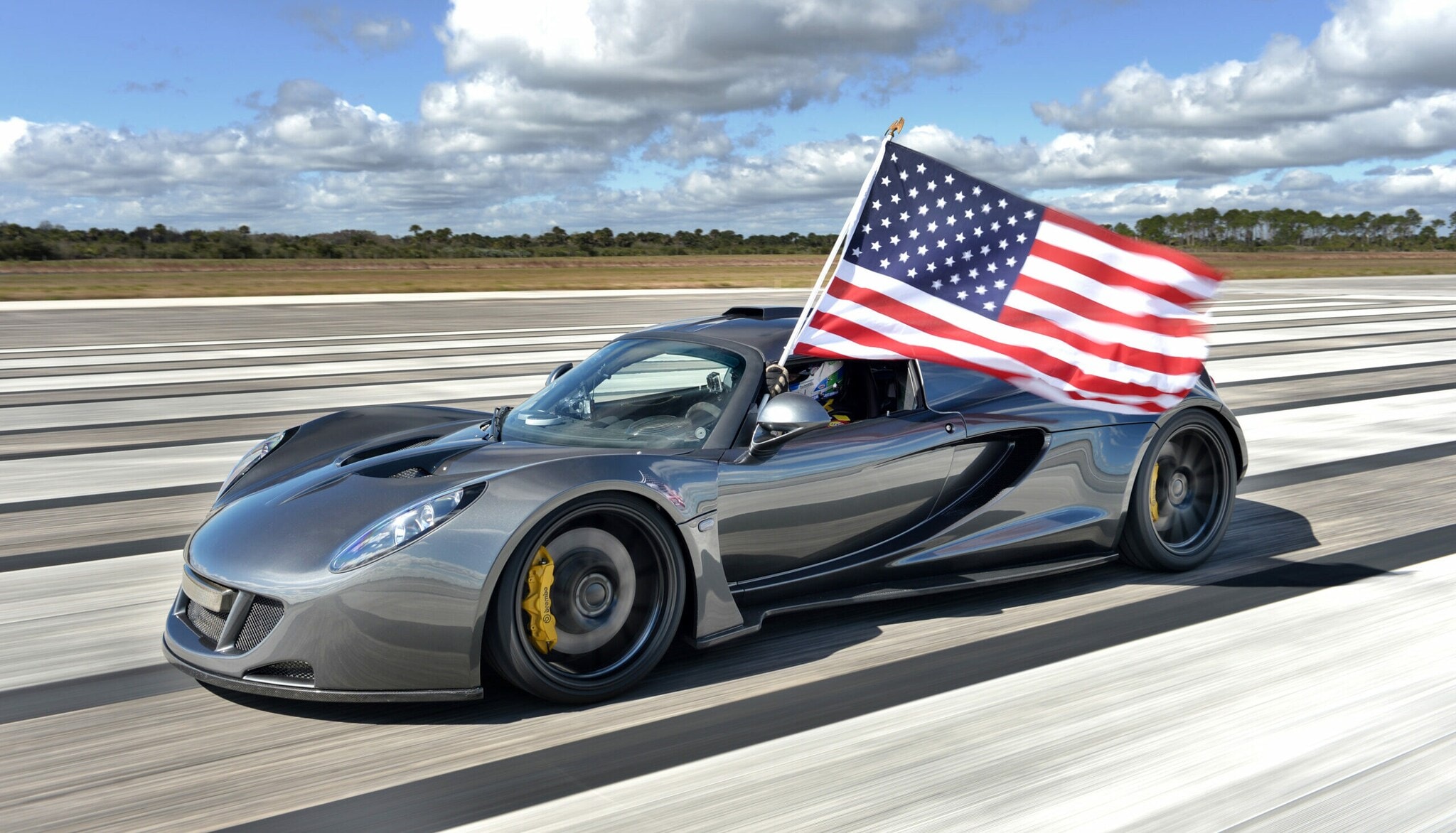 Der Hennessey Venom GT auf einer Rennstrecke, aus dem Fenster wird eine amerikanische Flagge gehalten. Der Hennessey Venom GT auf einer Rennstrecke, aus dem Fenster wird eine amerikanische Flagge gehalten.