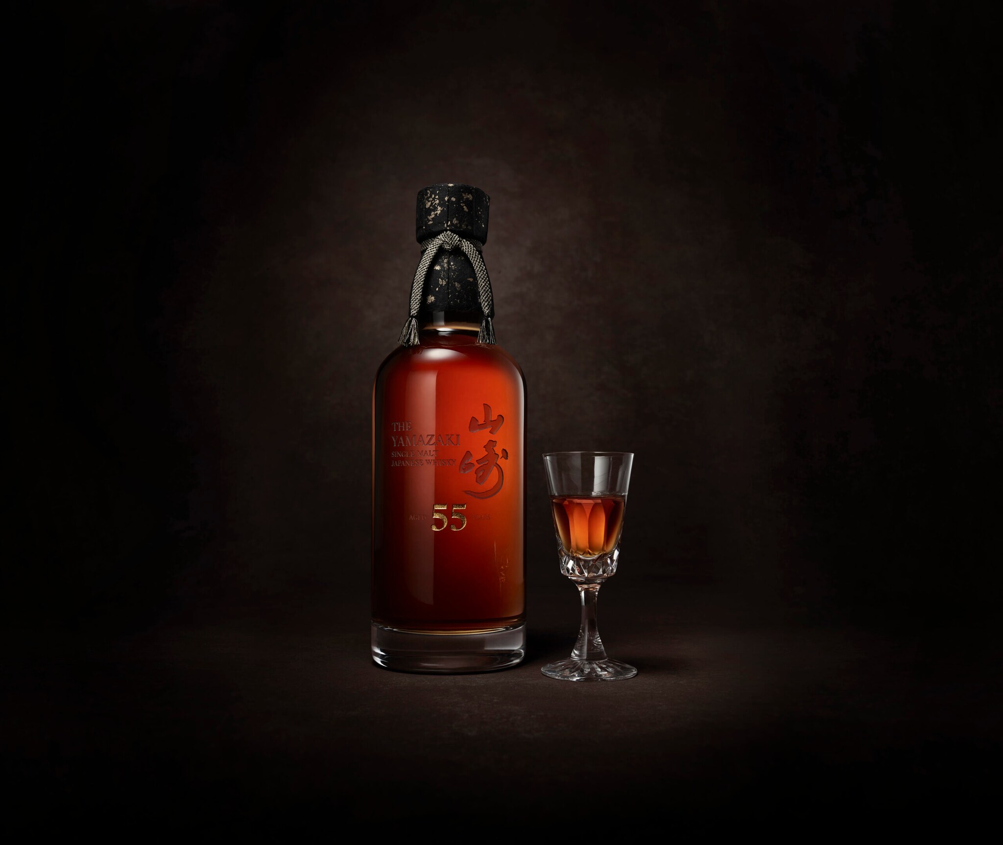 Eine Flasche des Whiskys Yamazaki 55 mit schwarzem Verschluss neben einem hochstieligen Whiskey-Glas vor schwarzem Hintergrund