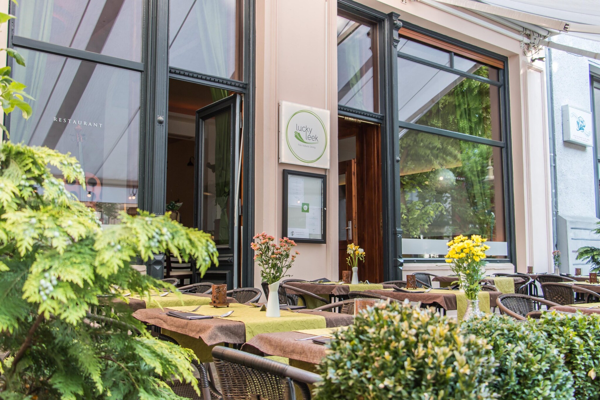 Eingang eines Restaurants mit Esstischen und Begrünung im Außenbereich