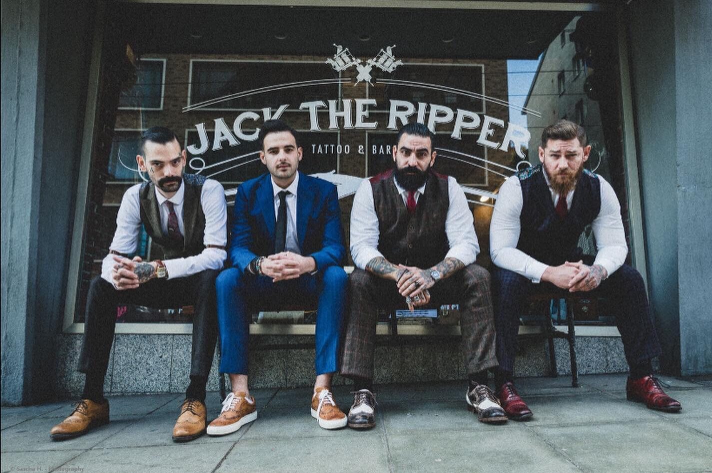 Vier Barbiere in klassischer Herrenkleidung sitzen vor einem Barbershop