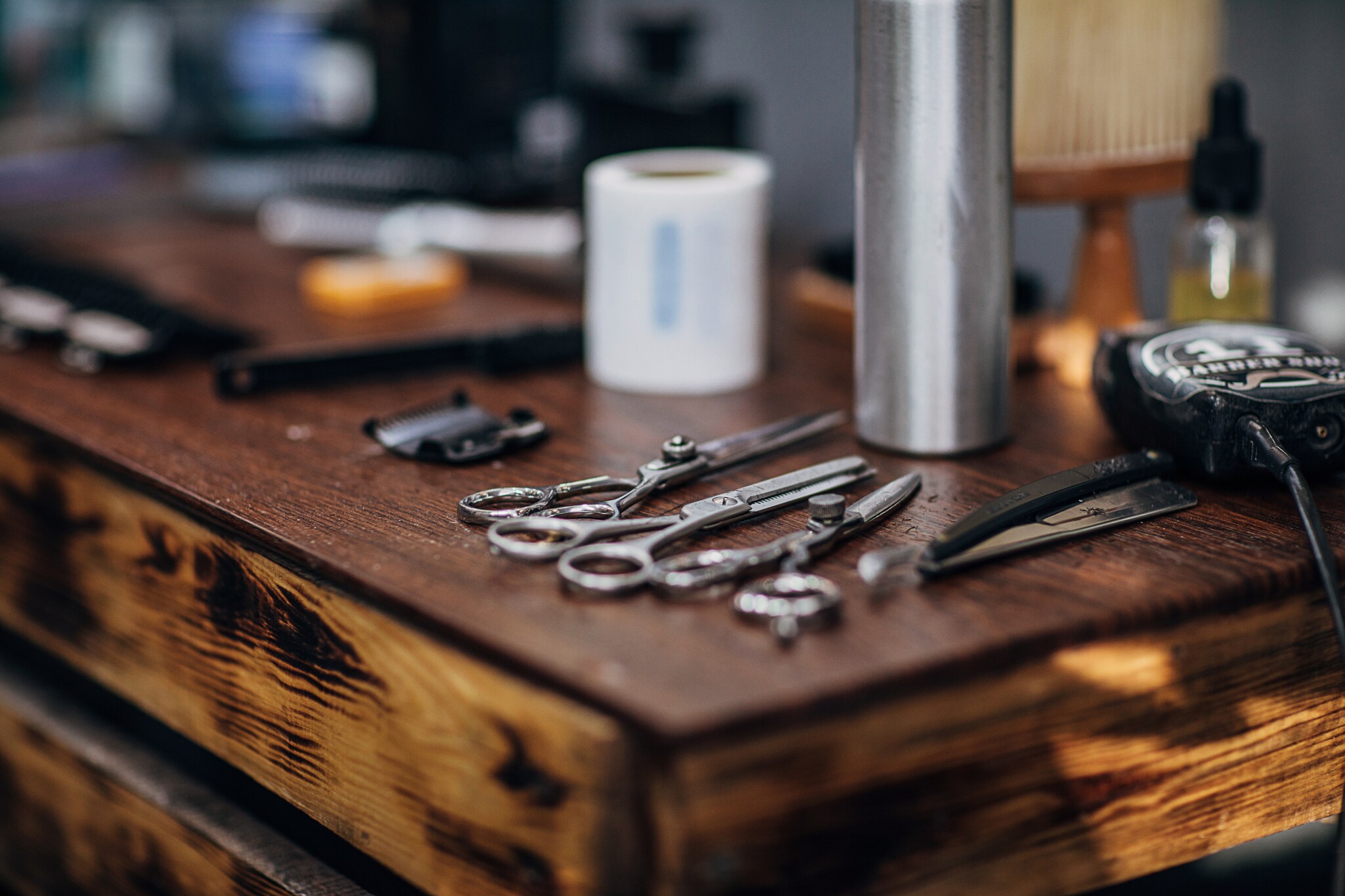 Barber-Zubehör wie drei verschiedene Scheren, Rasiermesser, elektrischer Trimmer und eine silberne Flasche auf einem Holztisch