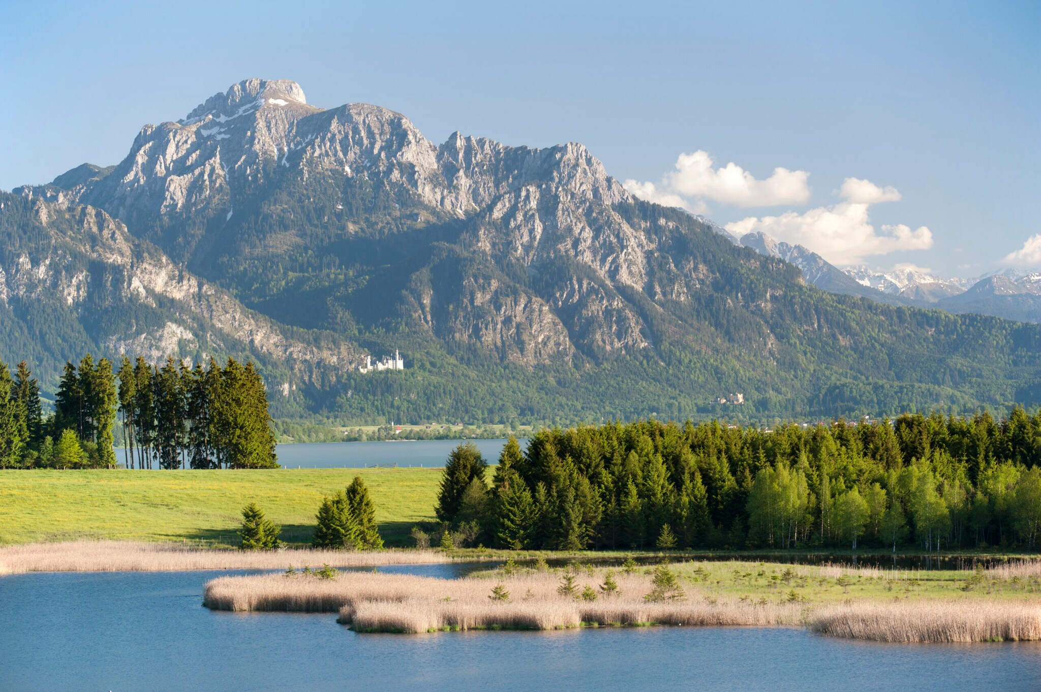 Ein See mit Wald vor Bergpanorama, im Hintergrund ein Schloss Ein See mit Wald vor Bergpanorama, im Hintergrund ein Schloss