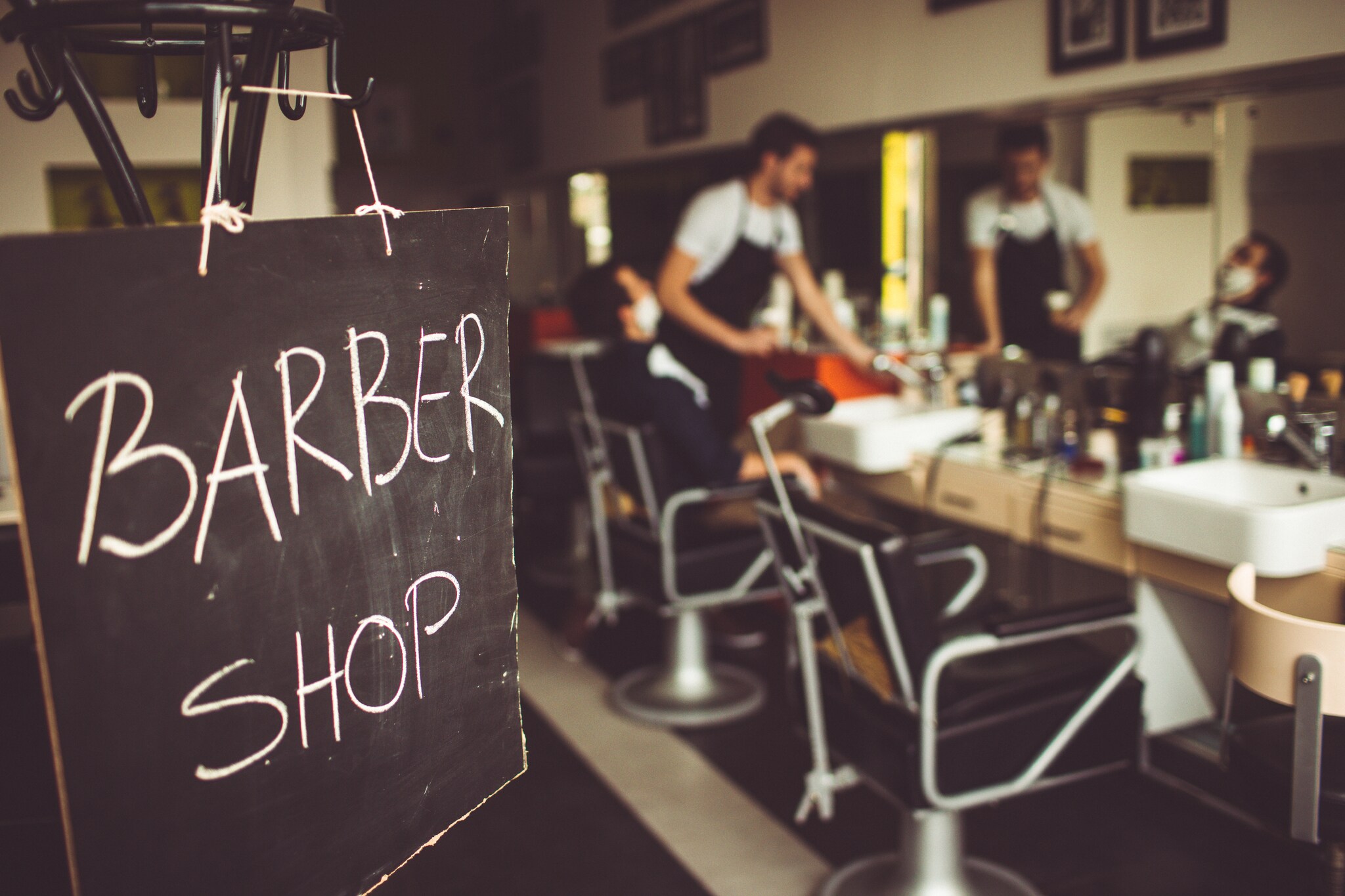 Eine Tafel auf der mit Kreide Barbershop steht, im Hintergrund ist ein Barbier mit seinem Kunden im Laden zu sehen