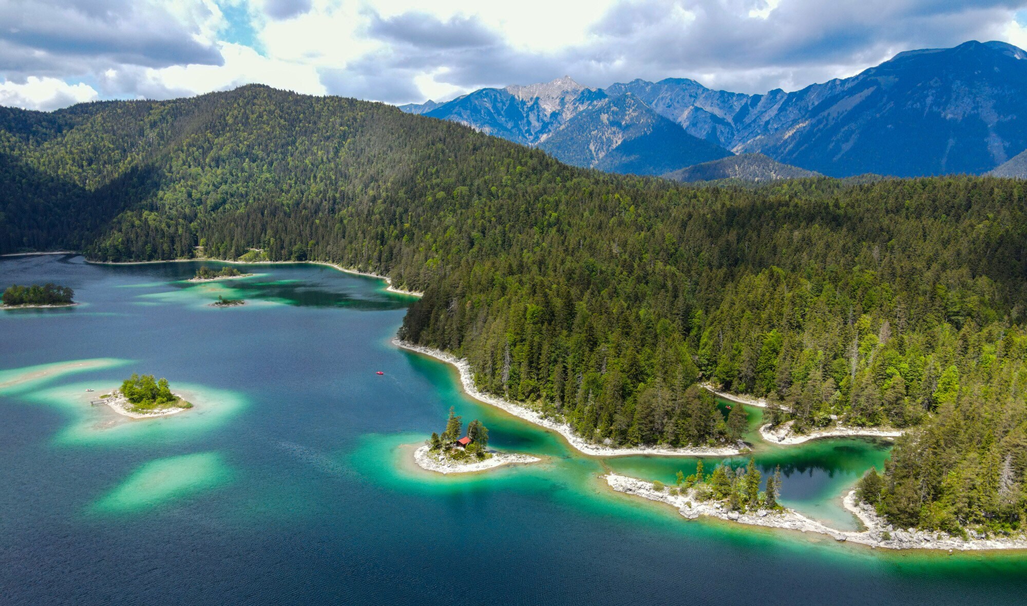Aufsicht auf einen See mit türkisblauem Wasser und vorgelagerten Inseln inmitten üppiger Waldlandschaft, im Hintergrund Bergpanorama