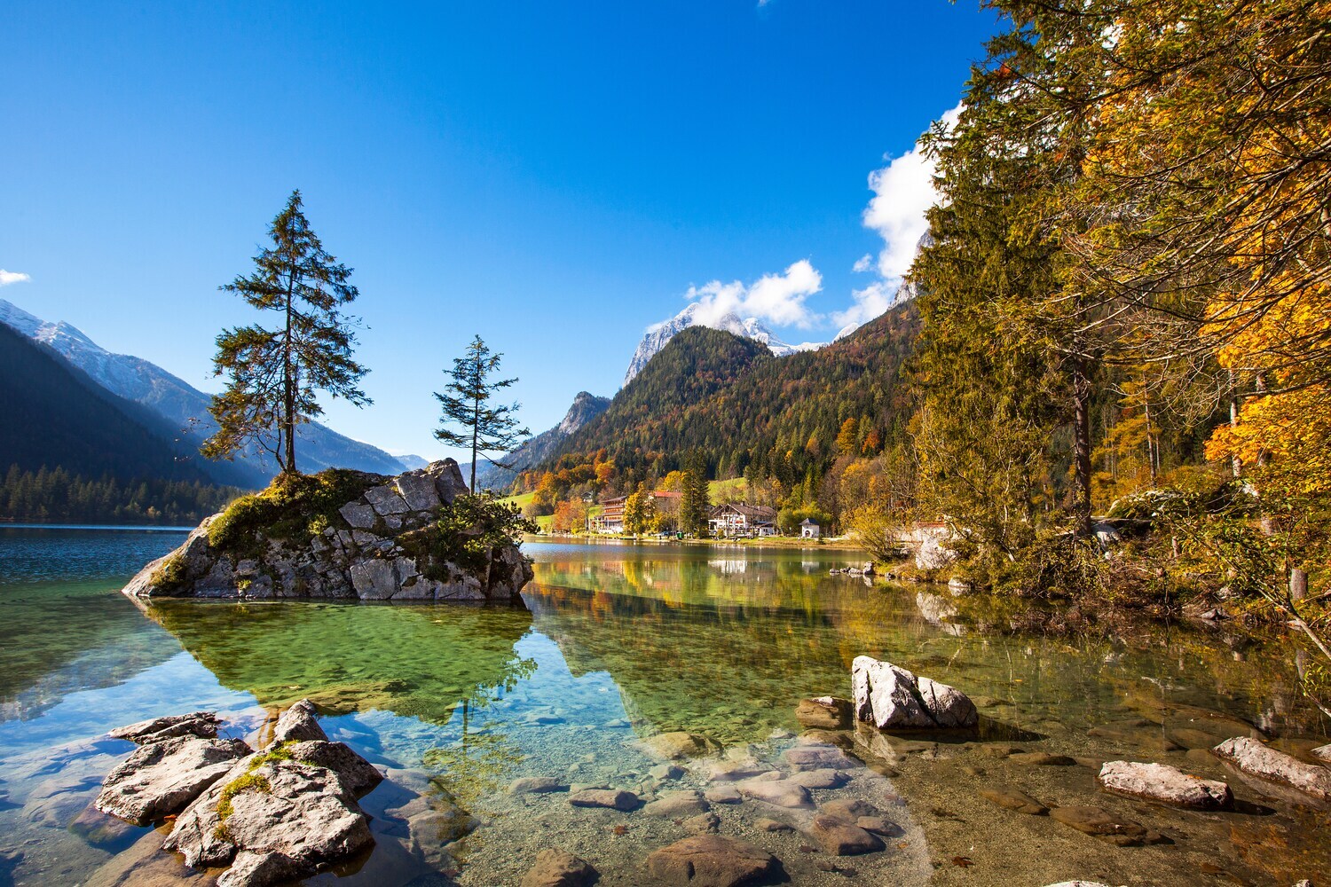 Ein See mit glasklarem Wasser mit Steinen, umgeben von herbstlichem Wald, im Hintergrund Bergpanorama Ein See mit glasklarem Wasser mit Steinen, umgeben von herbstlichem Wald, im Hintergrund Bergpanorama