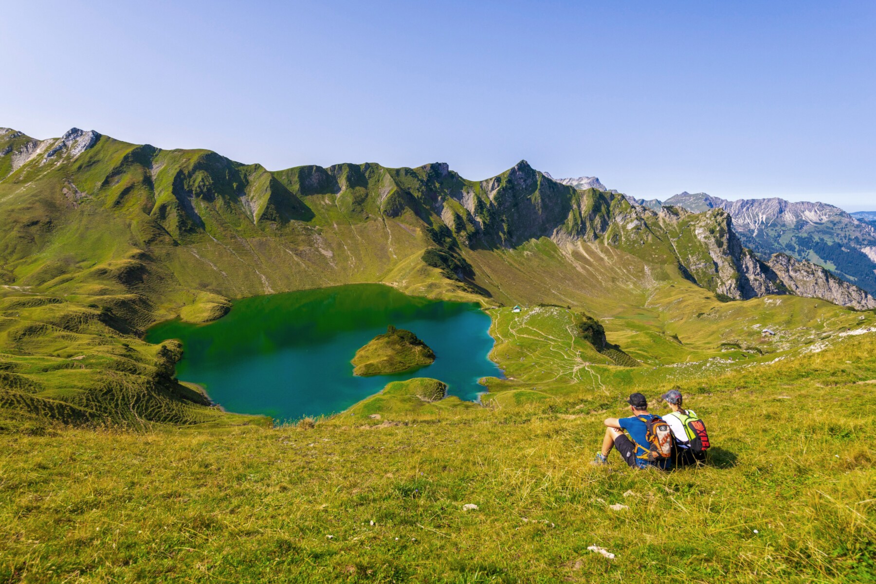 Zwei Wandernde sitzen mit Rucksäcken auf der Wiese vor einem Bergsee