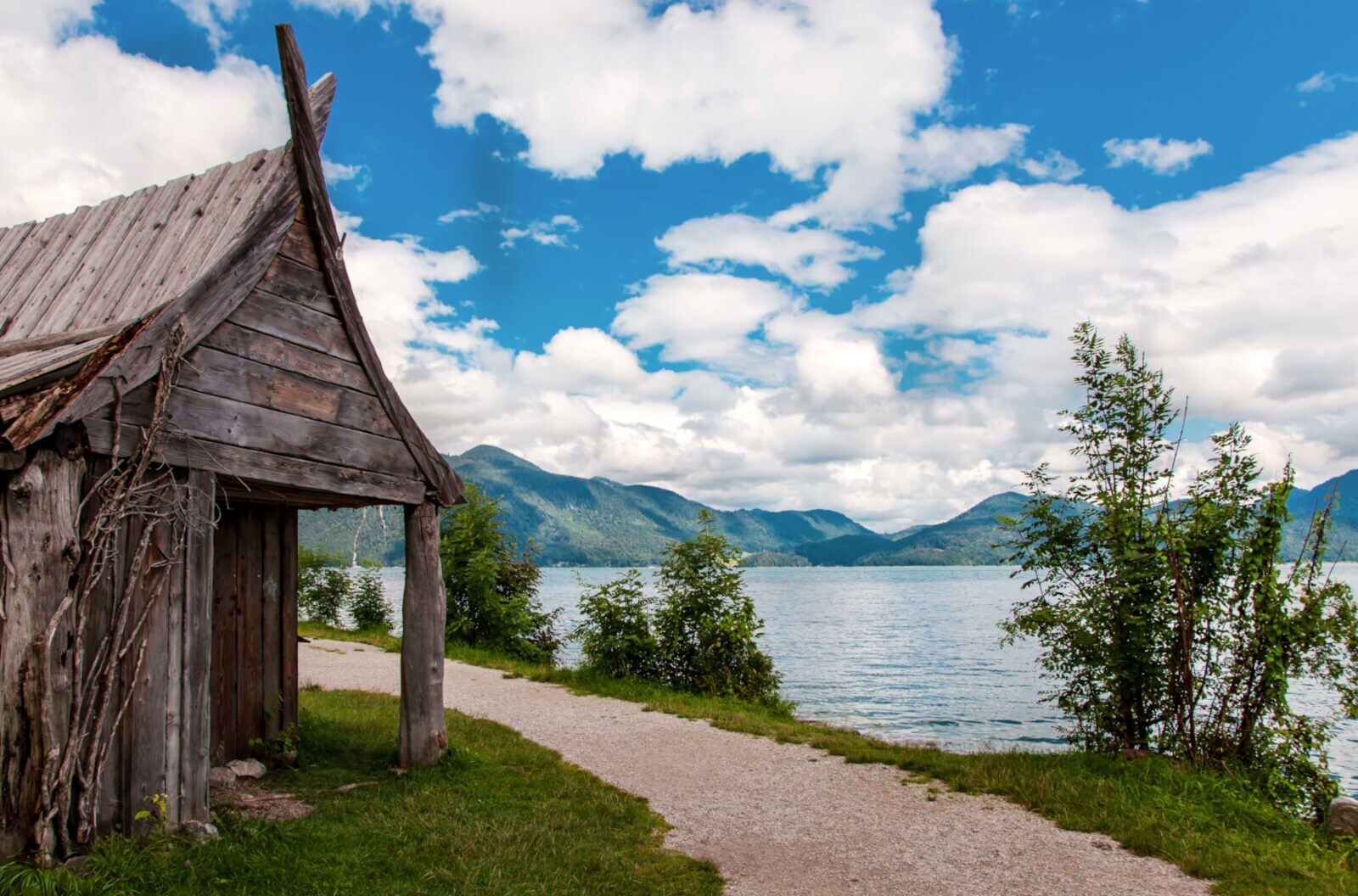 Ein Holzhaus an einem Weg am Ufer eines Sees mit Bergen im Hintergrund Ein Holzhaus an einem Weg am Ufer eines Sees mit Bergen im Hintergrund