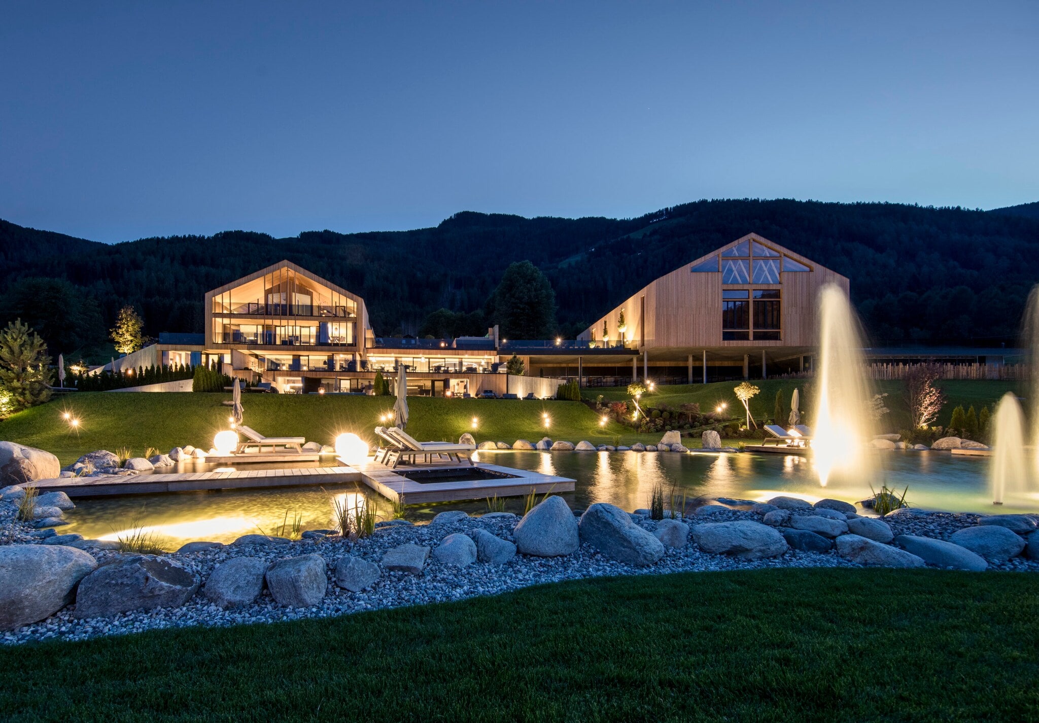 Eine luxuriöse, beleuchtete Hotelanlage aus Holzgebäuden mit Landschaftsgarten und Badeteich vor Bergpanorama bei Nacht Eine luxuriöse, beleuchtete Hotelanlage aus Holzgebäuden mit Landschaftsgarten und Badeteich vor Bergpanorama bei Nacht