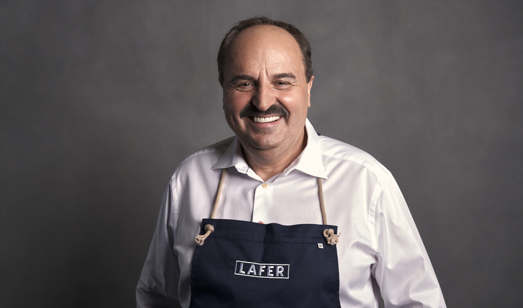 Studioporträt von Starkoch Johann Lafer, der in weißem Hemd und Schürze lächelnd in die Kamera blickt