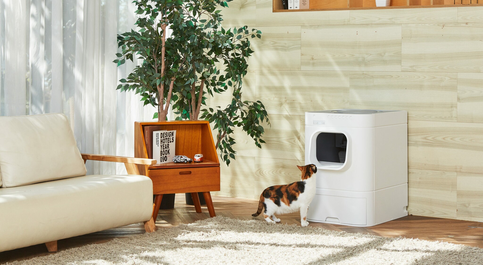 Eine etwa Waschmaschinen-große selbstreinigende weiße Katzentoilette, vor der eine rot-schwarz gefleckte Katze in einem skandinavisch eingerichteten Wohnzimmer steht