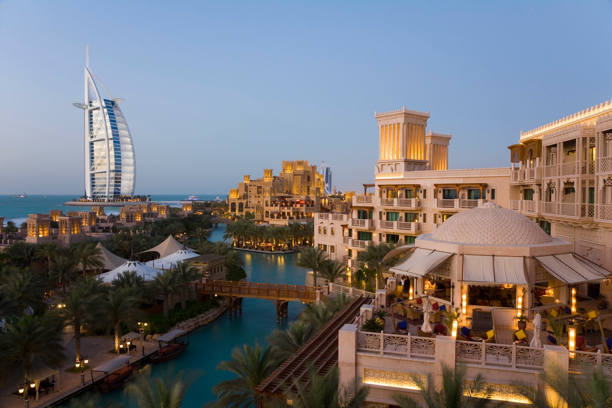 Stadtteil von Dubai mit Gebäuden an einem Fluss in der Abenddämmerung, im Hintergrund das Hotel Burj Al Arab in Segelform am Wasser Stadtteil von Dubai mit Gebäuden an einem Fluss in der Abenddämmerung, im Hintergrund das Hotel Burj Al Arab in Segelform am Wasser