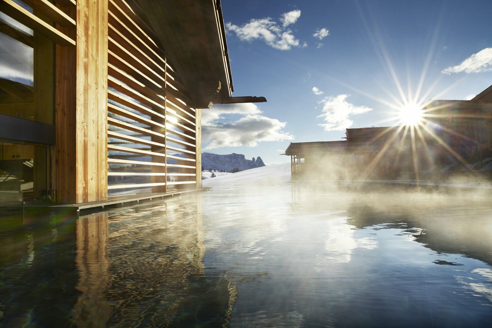 Ein Pool in der strahlenden Sonne, daneben Holzhäuser und Berge im Hintergrund Ein Pool in der strahlenden Sonne, daneben Holzhäuser und Berge im Hintergrund