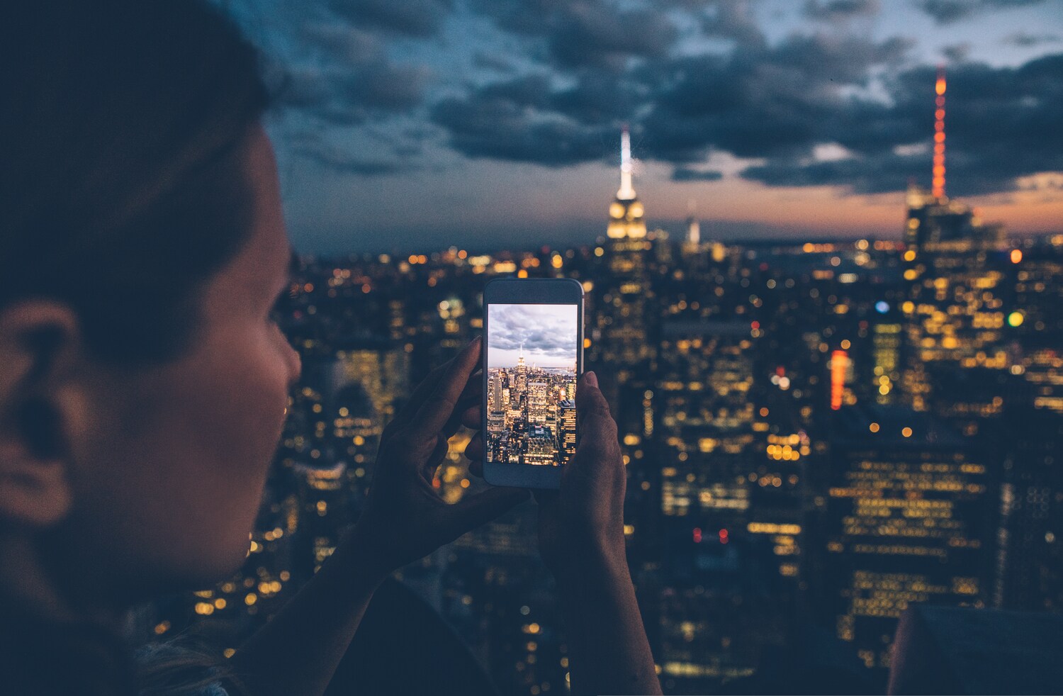 Eine Frau hält ein Smartphone in der Hand und fotografiert die nächtliche Skyline einer Stadt