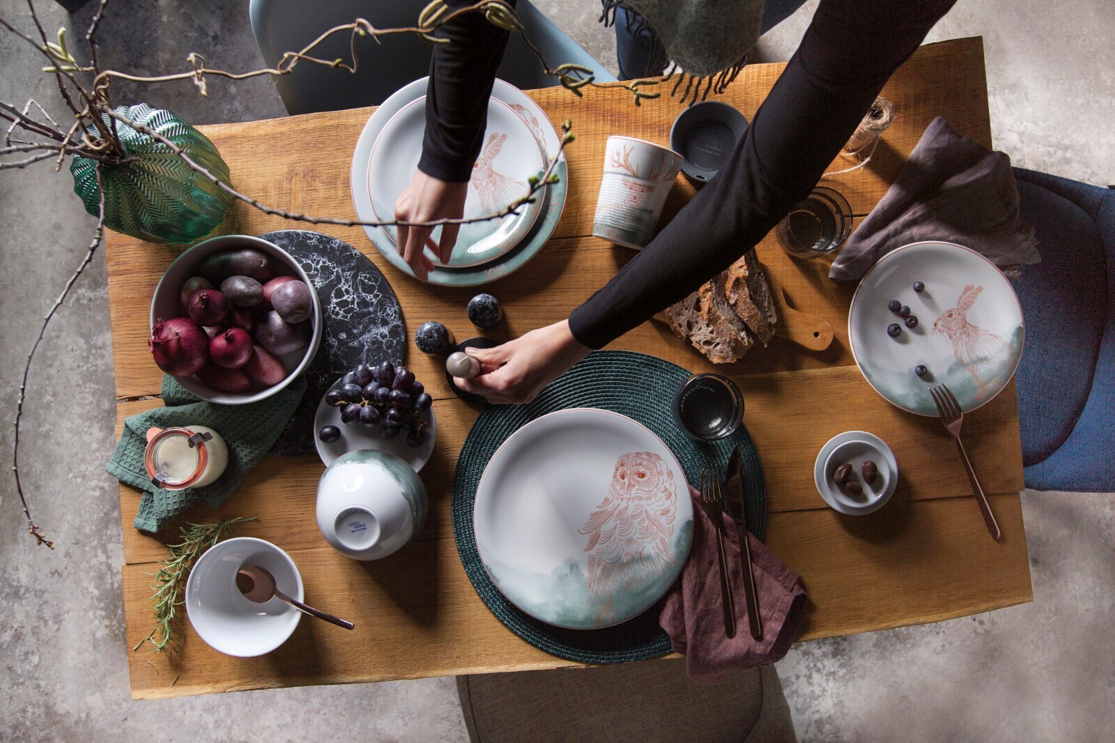 Ein gedeckter Holztisch mit kunstvoll bemaltem Porzellangeschirr, Brot, Obst und einer Vase mit Ästen