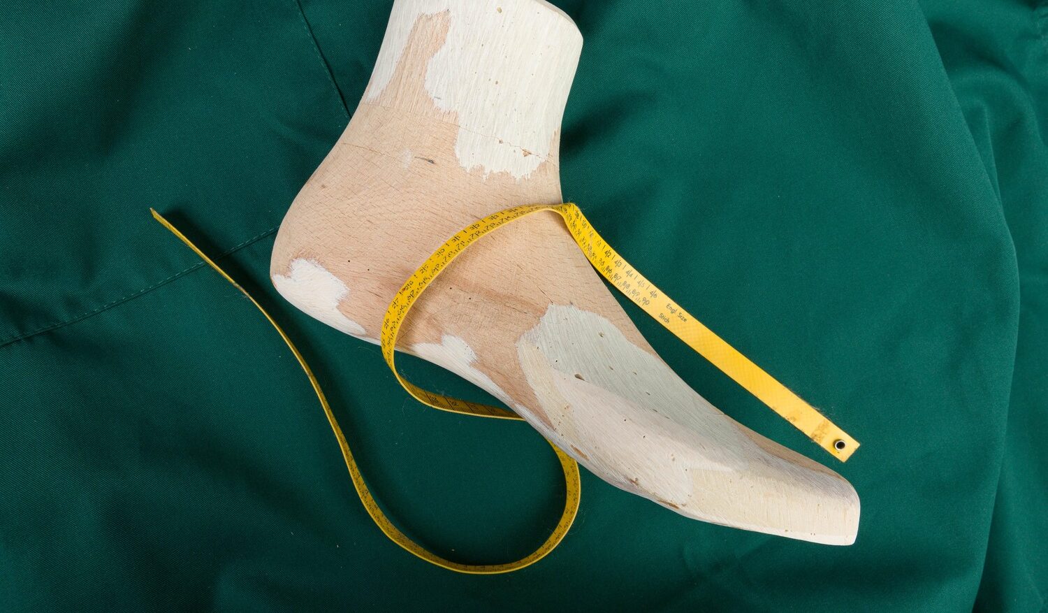 Ein hölzernes Schuh-Negativ liegt auf grünem Stoff, darüber ein gelbes Maßband.