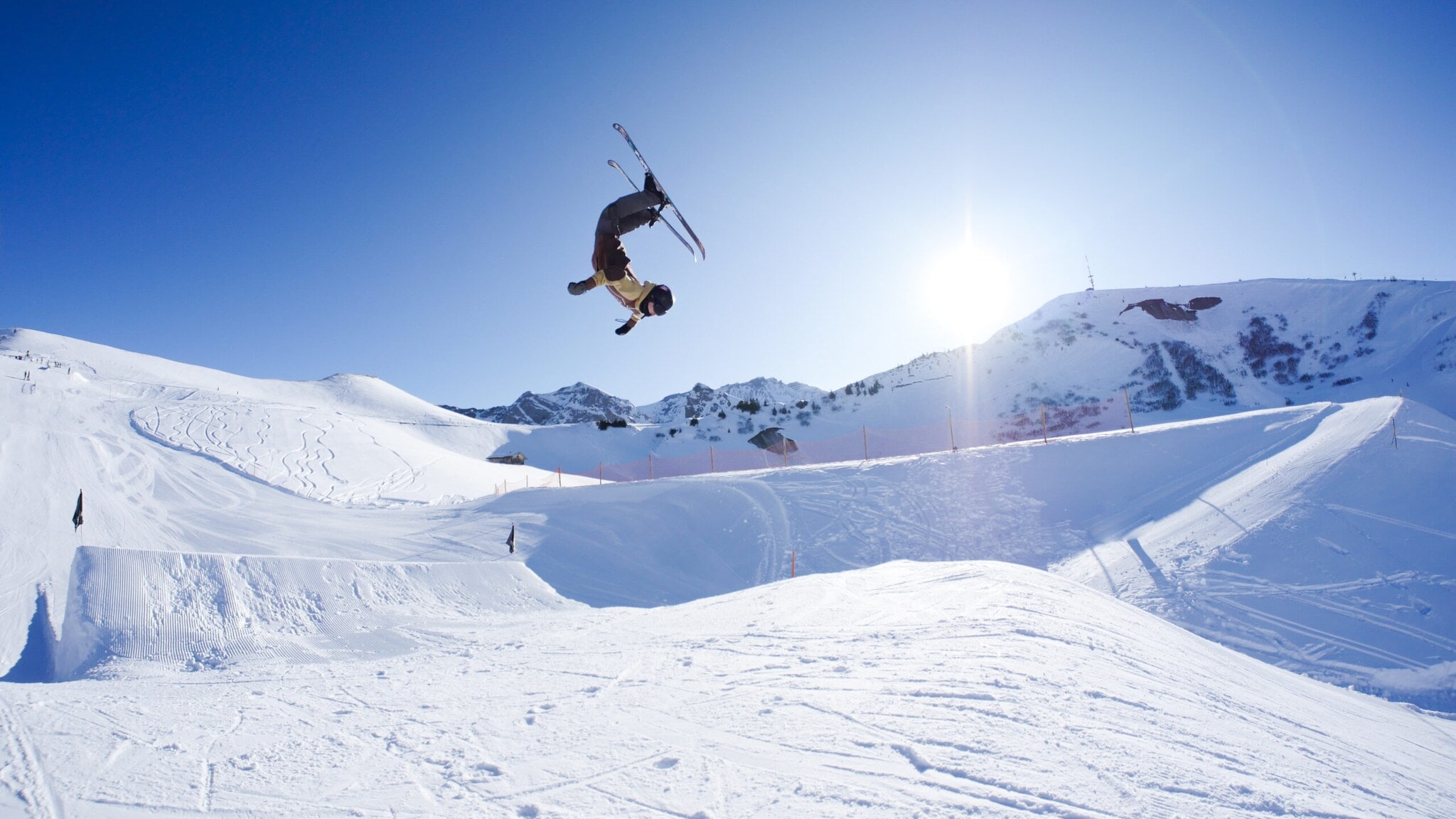 Eine Person, die mit Ski einen Salto in der Luft macht, im Hintergrund ein schneebedeckter Berg Eine Person, die mit Ski einen Salto in der Luft macht, im Hintergrund ein schneebedeckter Berg