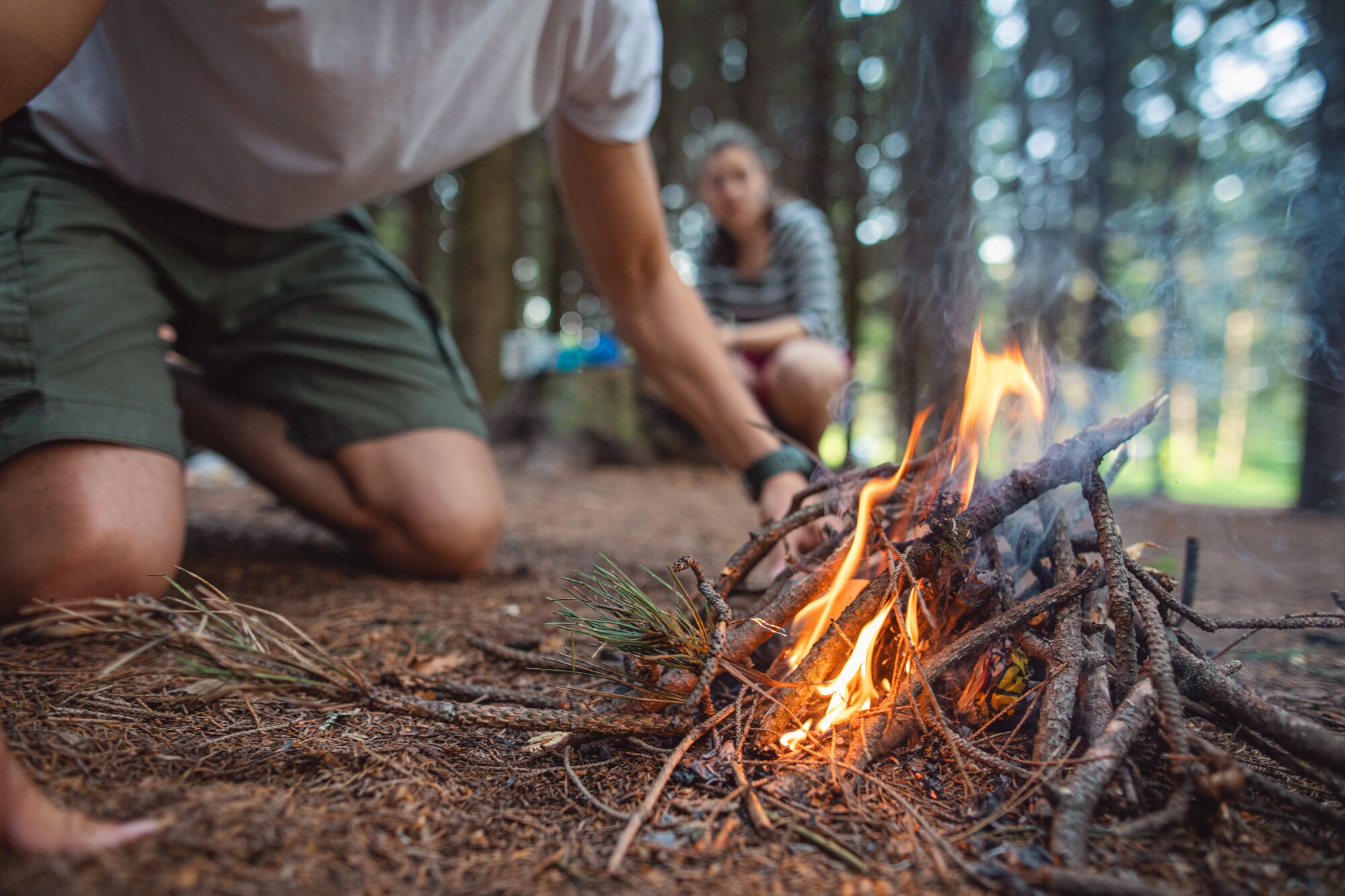 Zwei Personen hocken vor einer brennenden Feuerstelle aus Ästen in einem Wald