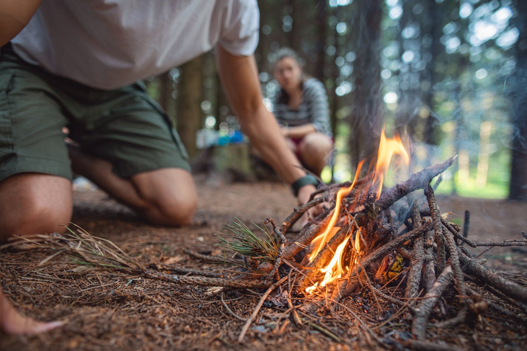 Zwei Personen hocken vor einer brennenden Feuerstelle aus Ästen in einem Wald Zwei Personen hocken vor einer brennenden Feuerstelle aus Ästen in einem Wald