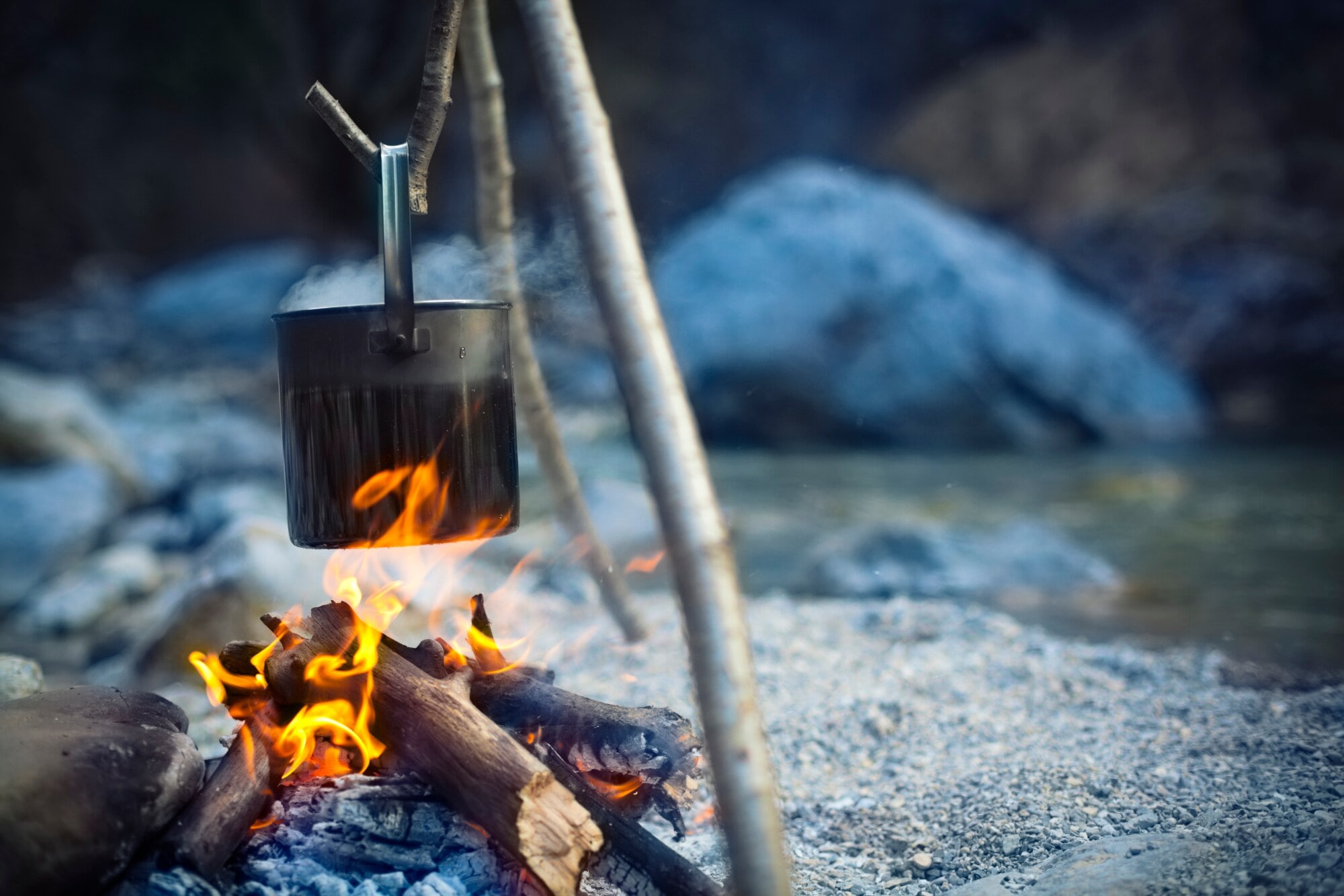 Ein Metalltopf hängt über einer Feuerstelle an einem See in der Dämmerung Ein Metalltopf hängt über einer Feuerstelle an einem See in der Dämmerung
