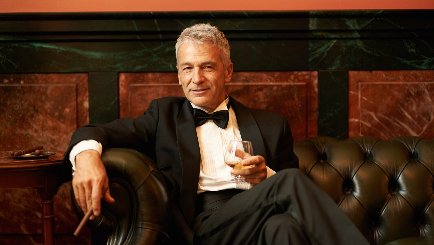 Ein Mann im Smoking mit Whiskyglas und Zigarre sitzt auf einem Ledersofa.