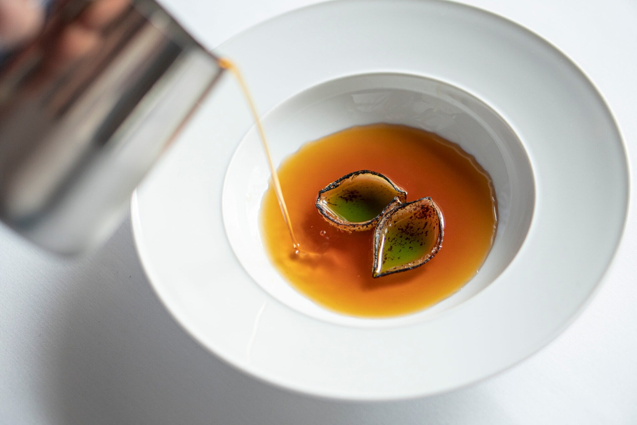 Aus einem Kännchen wird eine honiggelbe Flüssigkeit in einen weißen Teller mit zwei Blütenblättern gegossen