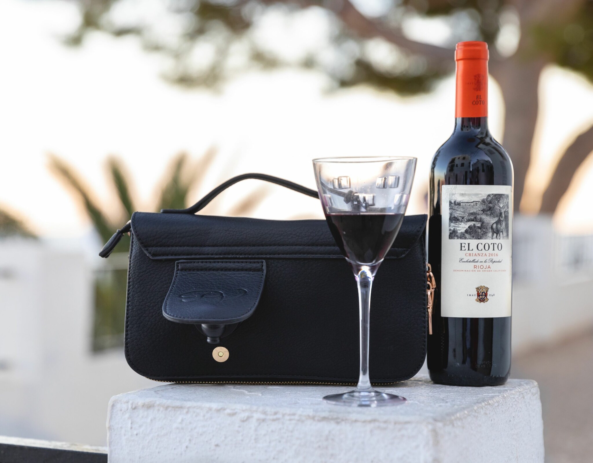 Eine Tasche mit kleiner Seitentasche inklusive Zapfhahn, daneben eine Weinflasche mit Glas