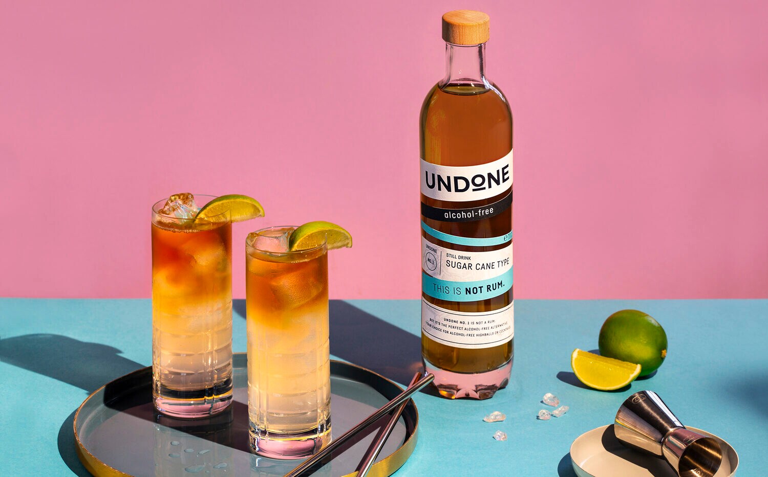 Zwei Gläser und eine Flasche mit Aufdruck auf einem Tisch, daneben Limetten und ein Barmaß