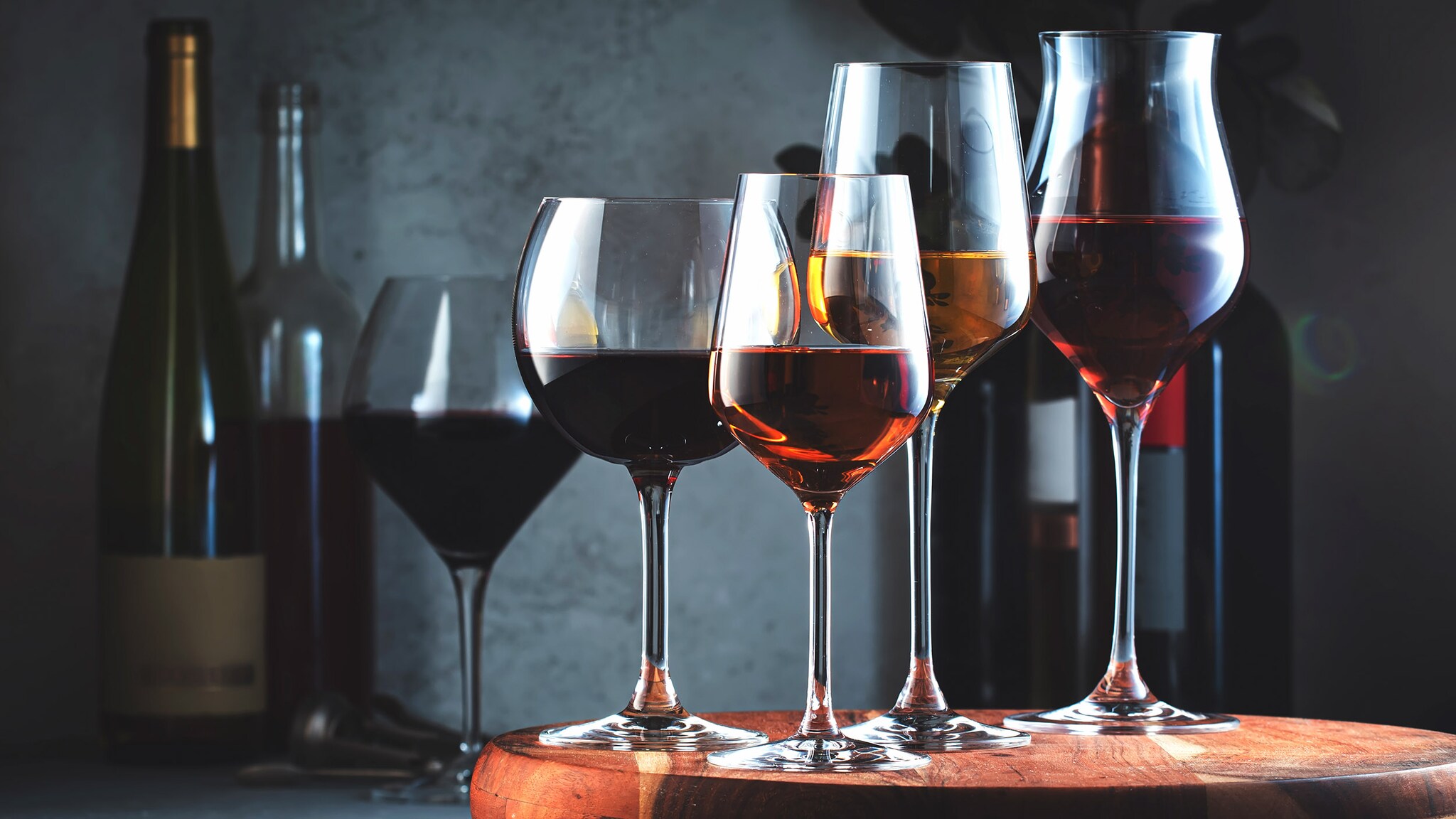 amex-magazin-hochwertige-weinglaeser Mehrere gefüllte Weingläser stehen nebeneinander auf einem Brett, dahinter Weinflaschen vor dunklem Hintergrund.