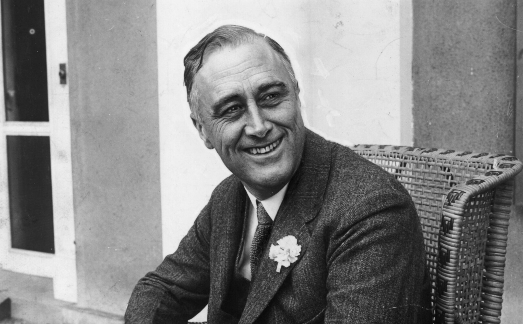 Eine Schwarzweiß-Aufnahme eines lachenden Mannes im Sakko, auf einem Korbstuhl sitzend