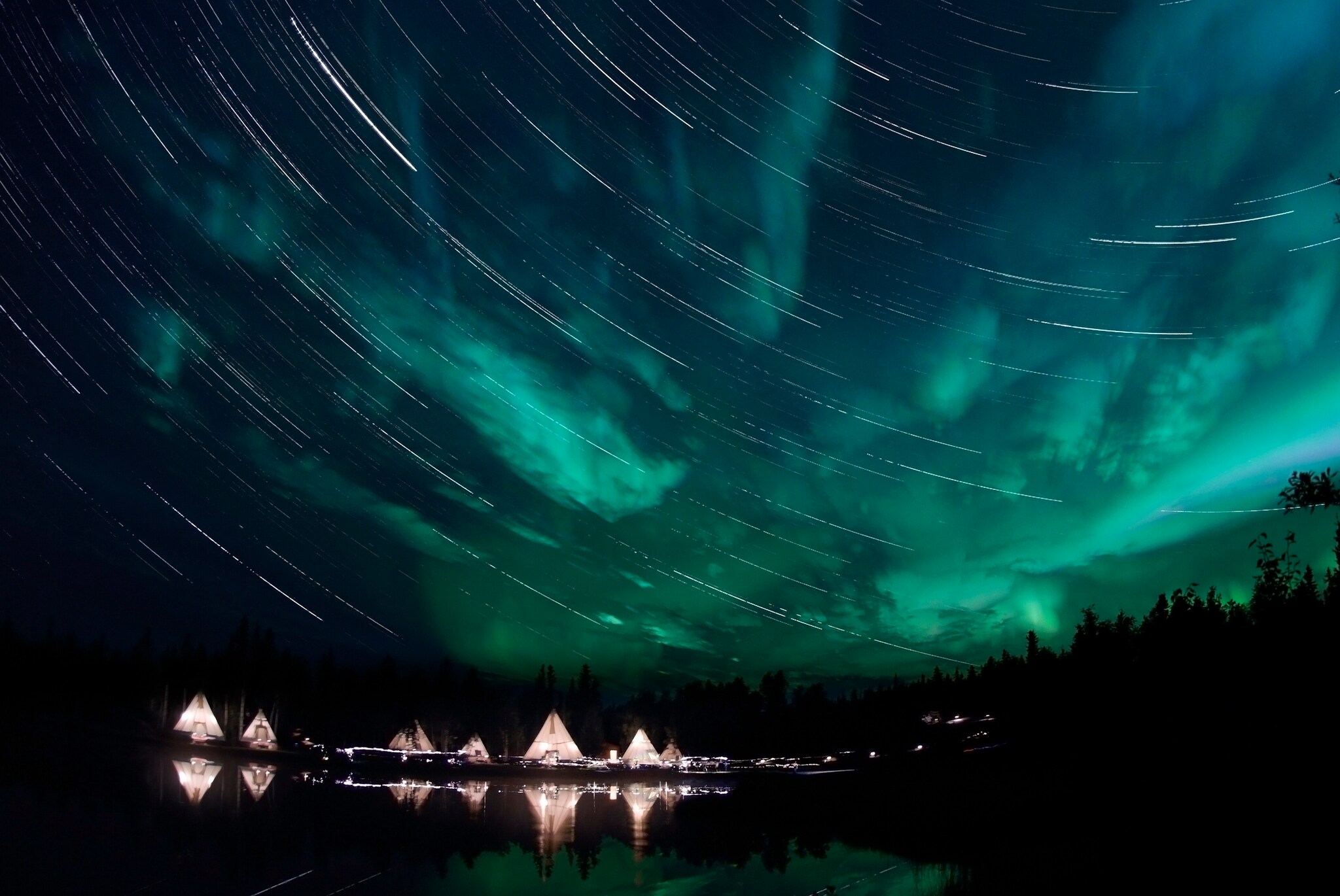 Grüne Polarlichter am Nachthimmel, unter ihnen ein beleuchtetes Tipi-Dorf