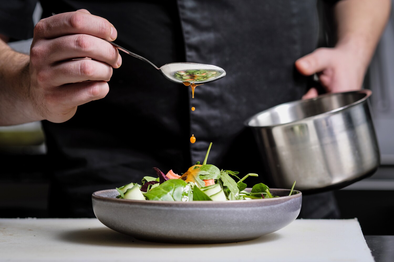 Nahaufnahme einer Hand, die mit einem Löffel gelbes Dressing über einen Teller mit Rohkost träufelt