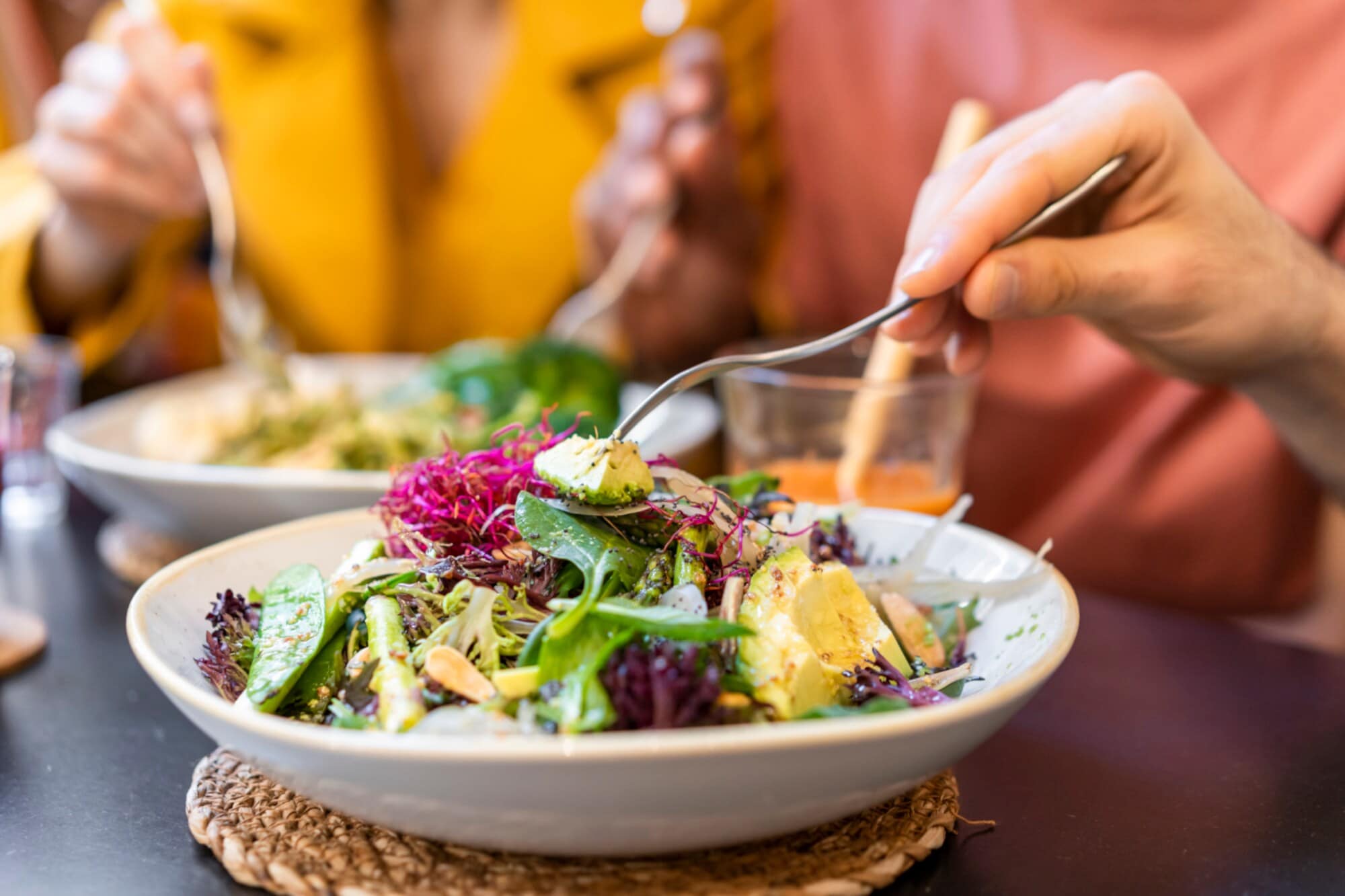 Nahaufnahme zweier weißer Teller mit vegetarischer Rohkost auf einem Tisch, drumherum Hände mit Gabeln in den Gerichten Nahaufnahme zweier weißer Teller mit vegetarischer Rohkost auf einem Tisch, drumherum Hände mit Gabeln in den Gerichten