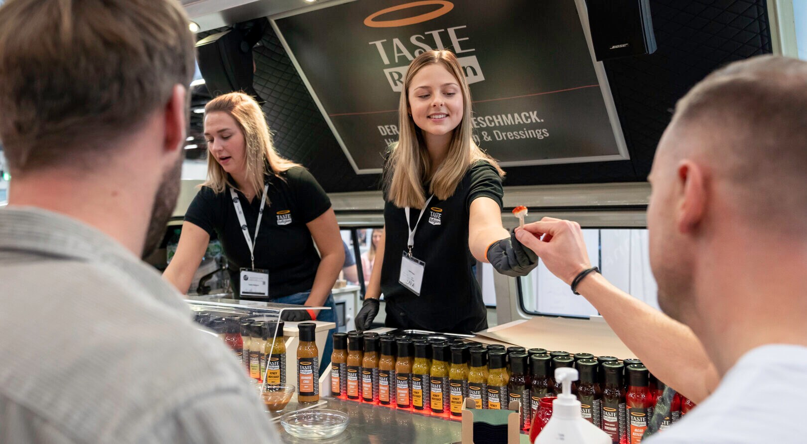 Einem Messebesucher wird ein Probierhäppchen von einer Dame hinter einer Theke gereicht Einem Messebesucher wird ein Probierhäppchen von einer Dame hinter einer Theke gereicht