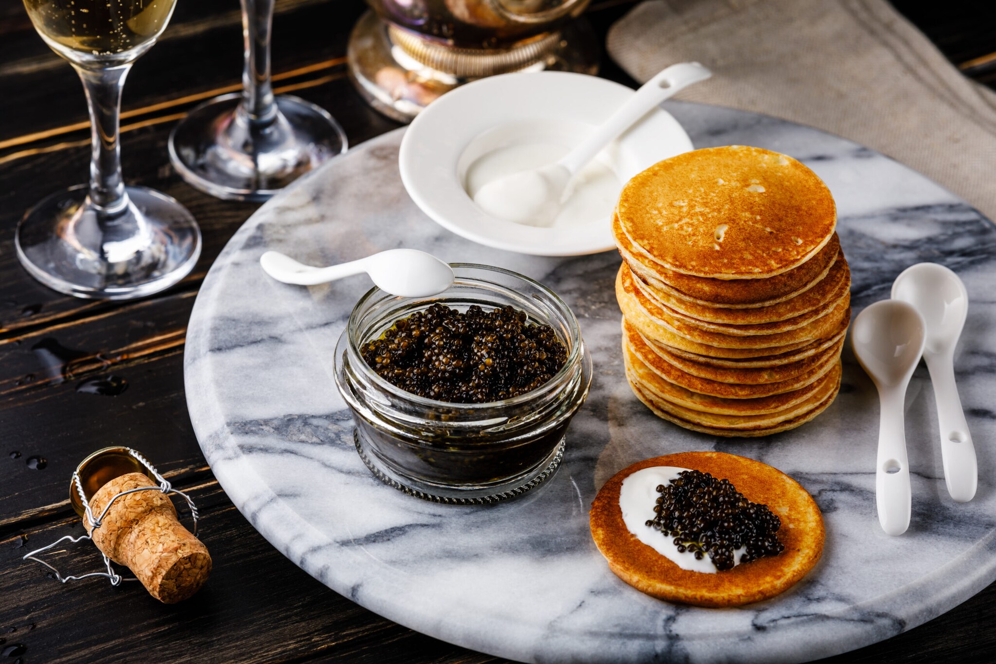 Kaviar im Glas, ein Stapel Blinis und Löffel auf einem Marmortisch, daneben Sektkorken und -gläser Kaviar im Glas, ein Stapel Blinis und Löffel auf einem Marmortisch, daneben Sektkorken und -gläser
