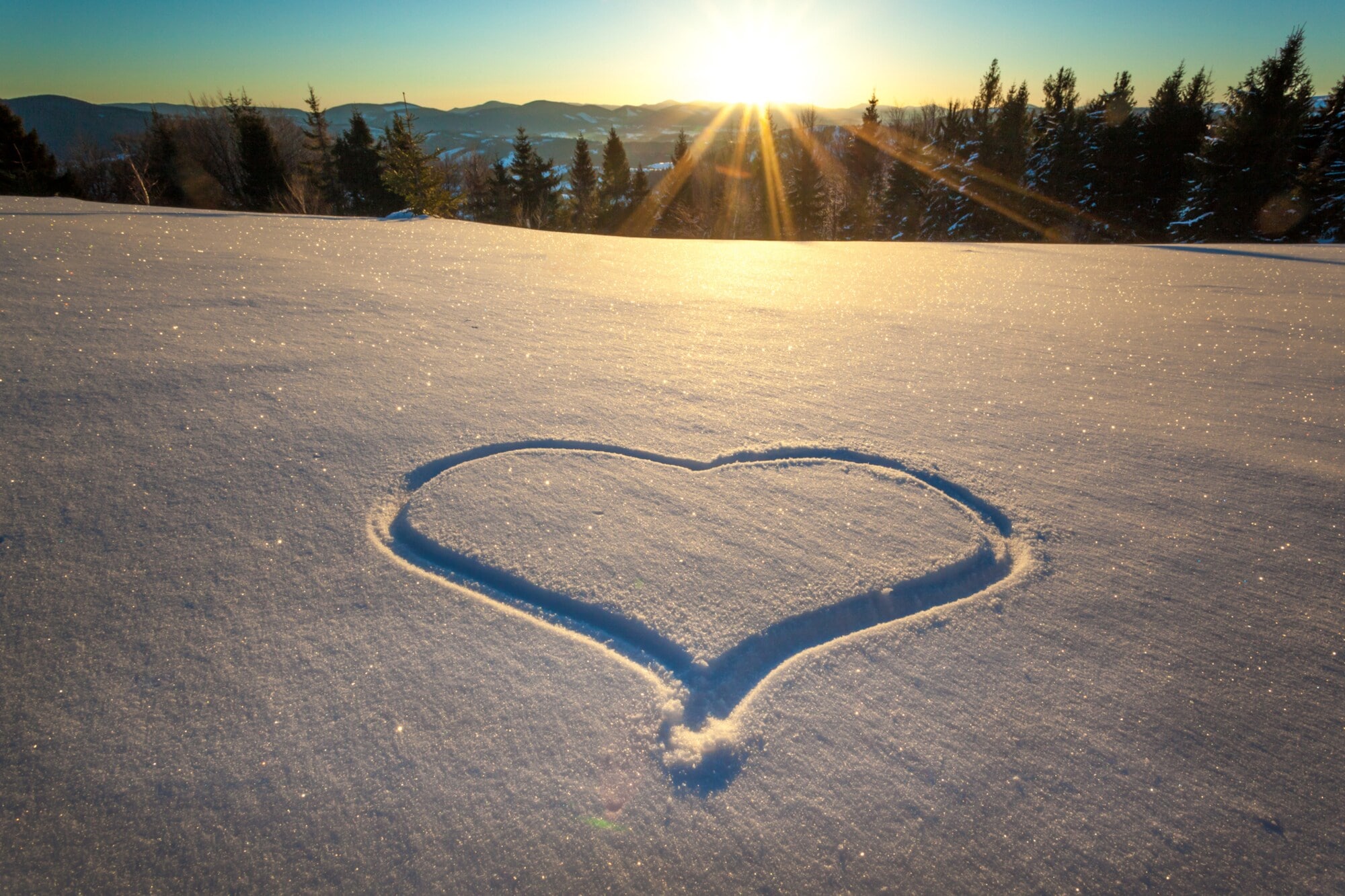 Ein Herz in den Schnee gezeichnet, im Hintergrund ein Wald und Berge Ein Herz in den Schnee gezeichnet, im Hintergrund ein Wald und Berge