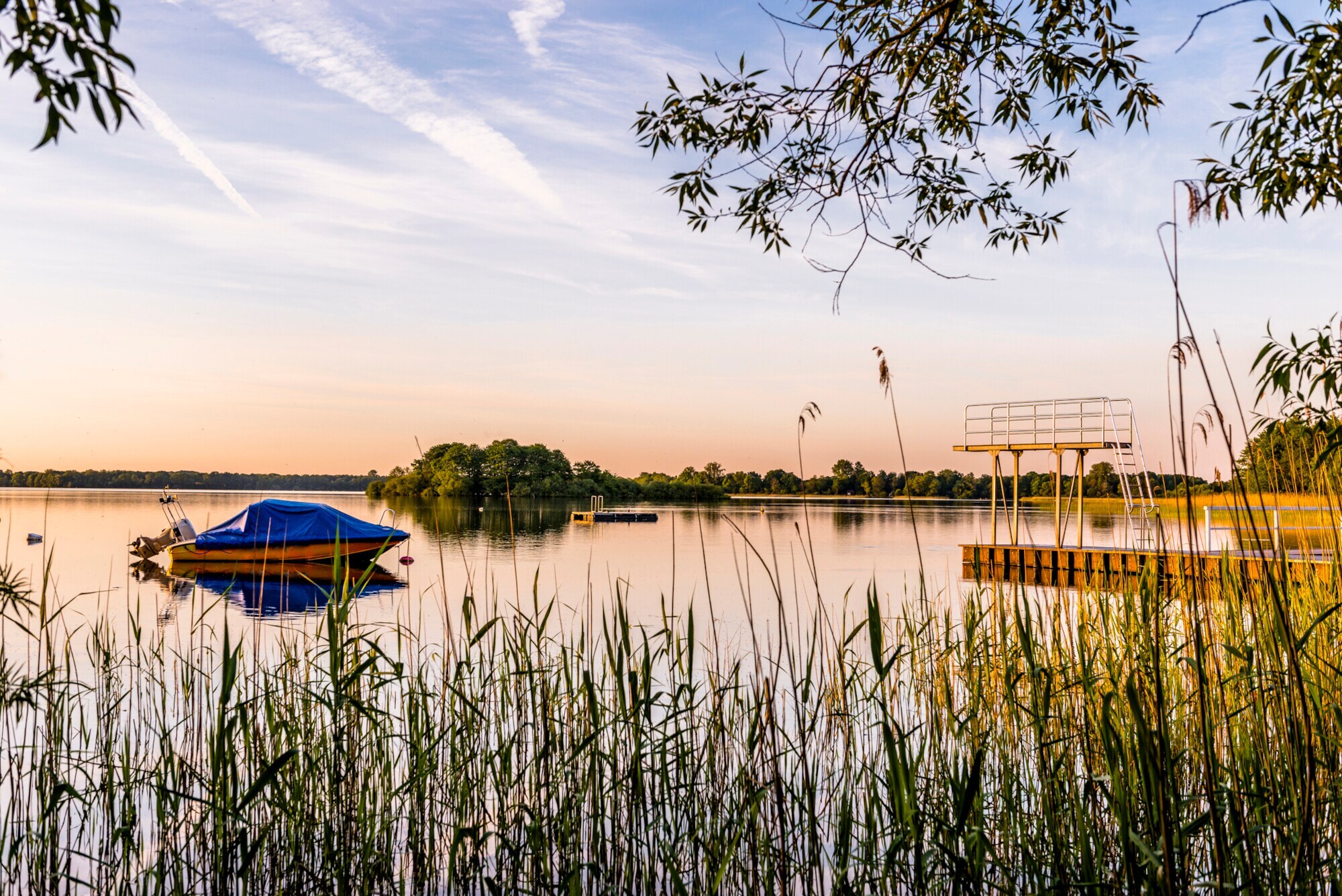 Ein See mit einem Boot und einem Sprungturm in der Abendsonne, im Vordergrund Schilf