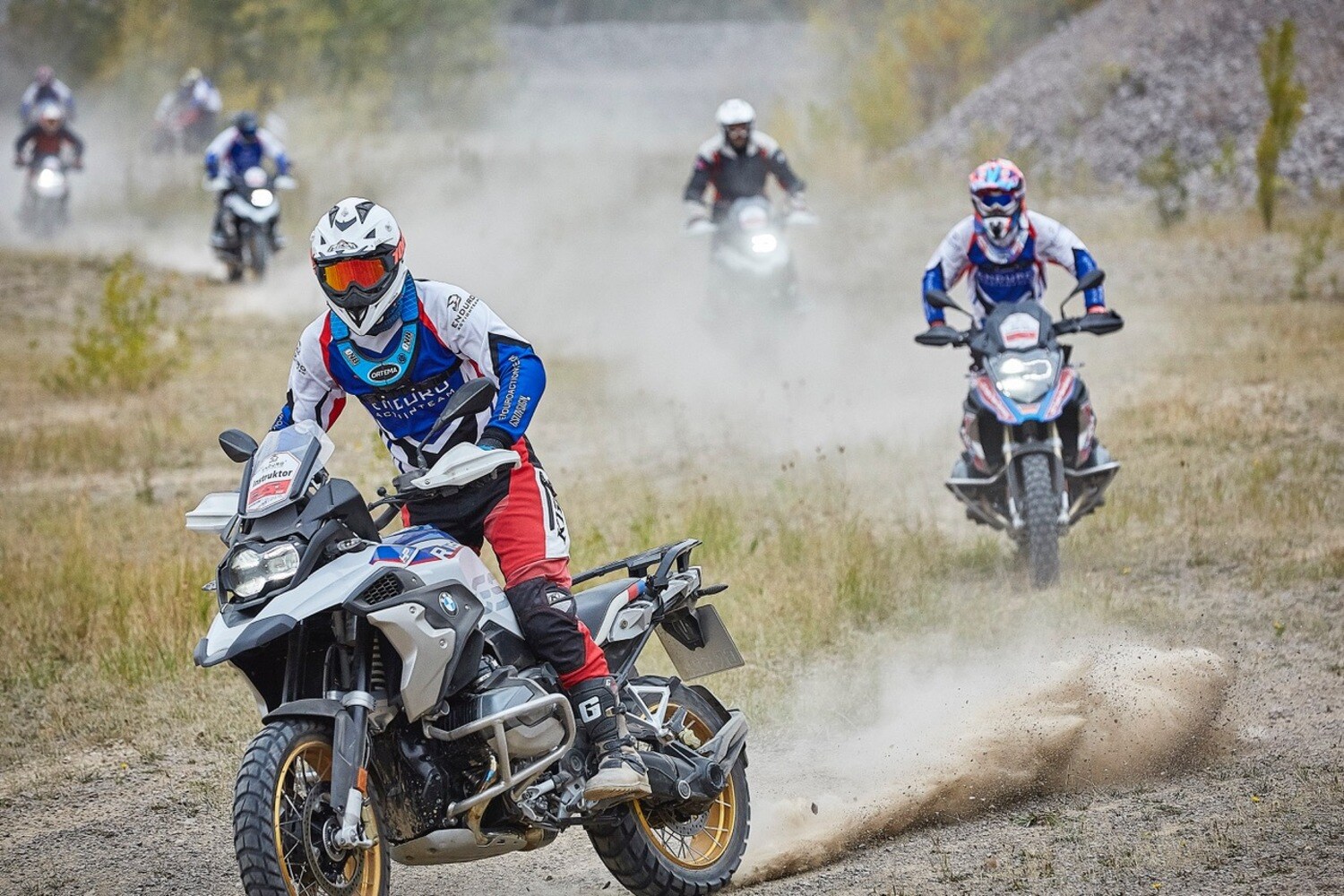 Eine Gruppe von Endurofahrer:innen fährt mit Motorrädern über eine Schotterfläche. Eine Gruppe von Endurofahrer:innen fährt mit Motorrädern über eine Schotterfläche.