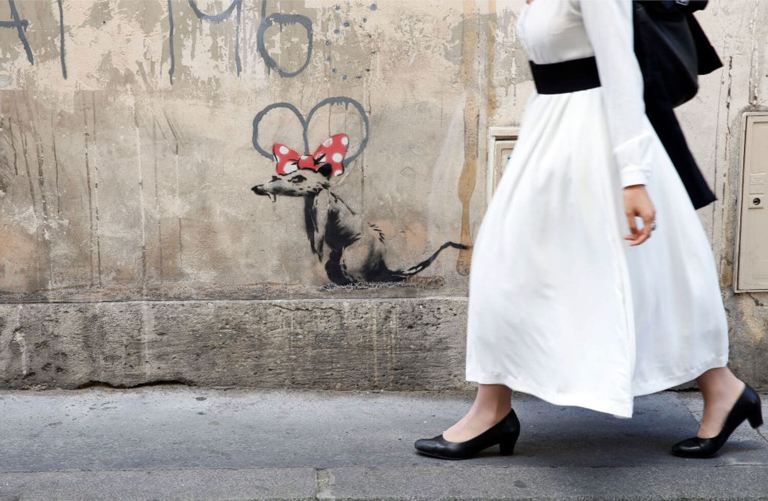 Eine Frau in langem, weißen Kleid läuft an einem Banksy-Werk vorbei: eine Ratte mit roter Minnie-Mouse-Schleife auf dem Kopf. Eine Frau in langem, weißen Kleid läuft an einem Banksy-Werk vorbei: eine Ratte mit roter Minnie-Mouse-Schleife auf dem Kopf.