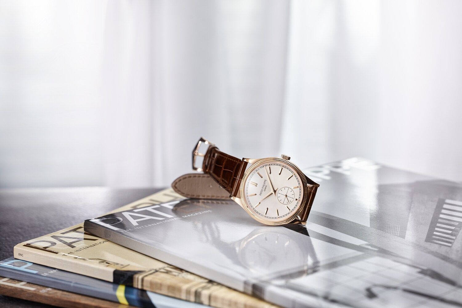 Eine klassische Herrenuhr von Patek Philippe mit braunem Lederarmband, goldener Lünette und hellem Zifferblatt auf Hochglanzmagazinen Eine klassische Herrenuhr von Patek Philippe mit braunem Lederarmband, goldener Lünette und hellem Zifferblatt auf Hochglanzmagazinen