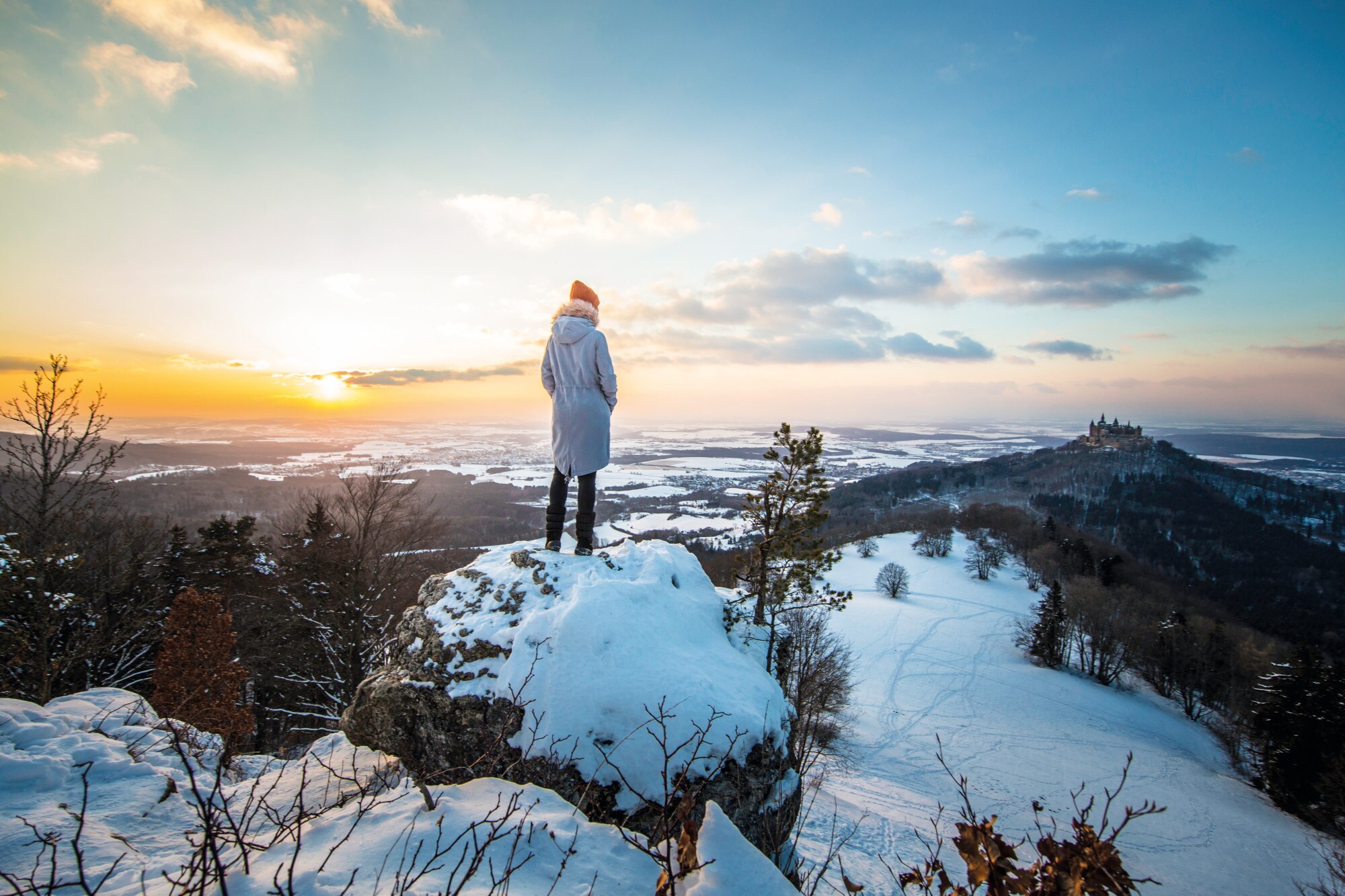 Rückansicht einer Person, die auf einem Felsvorsprung des Zeller Horns steht und auf die gegenüberliegende Burg Hohenzollern blickt