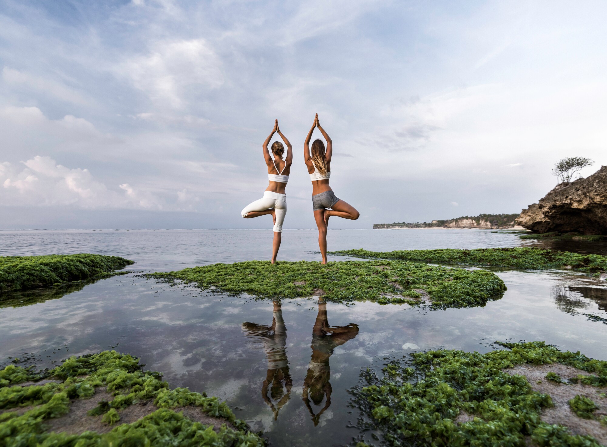 Zwei Frauen stehen am Wasser und machen zwei Yogaübungen