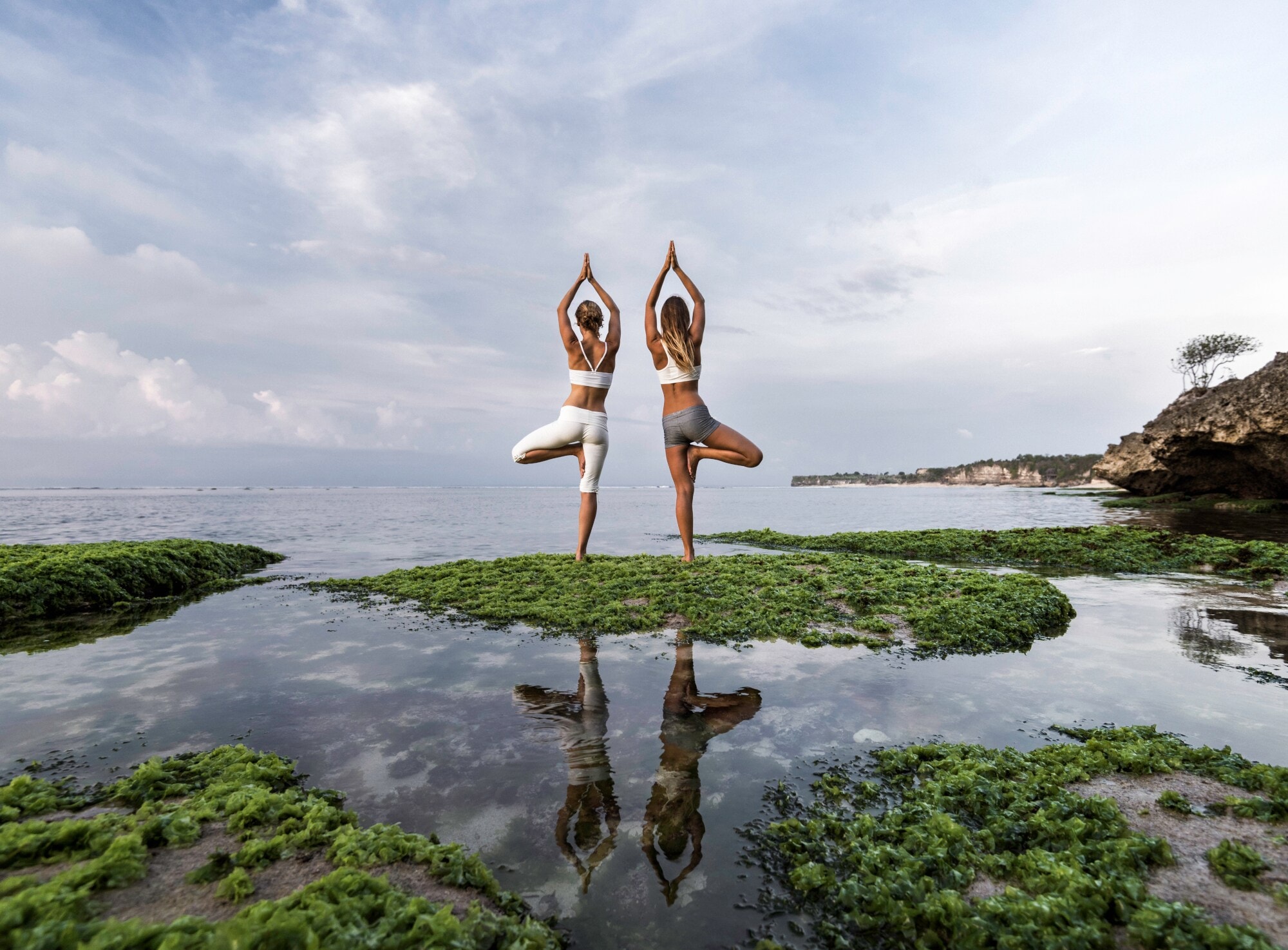 Zwei Frauen stehen am Wasser und machen zwei Yogaübungen Zwei Frauen stehen am Wasser und machen zwei Yogaübungen